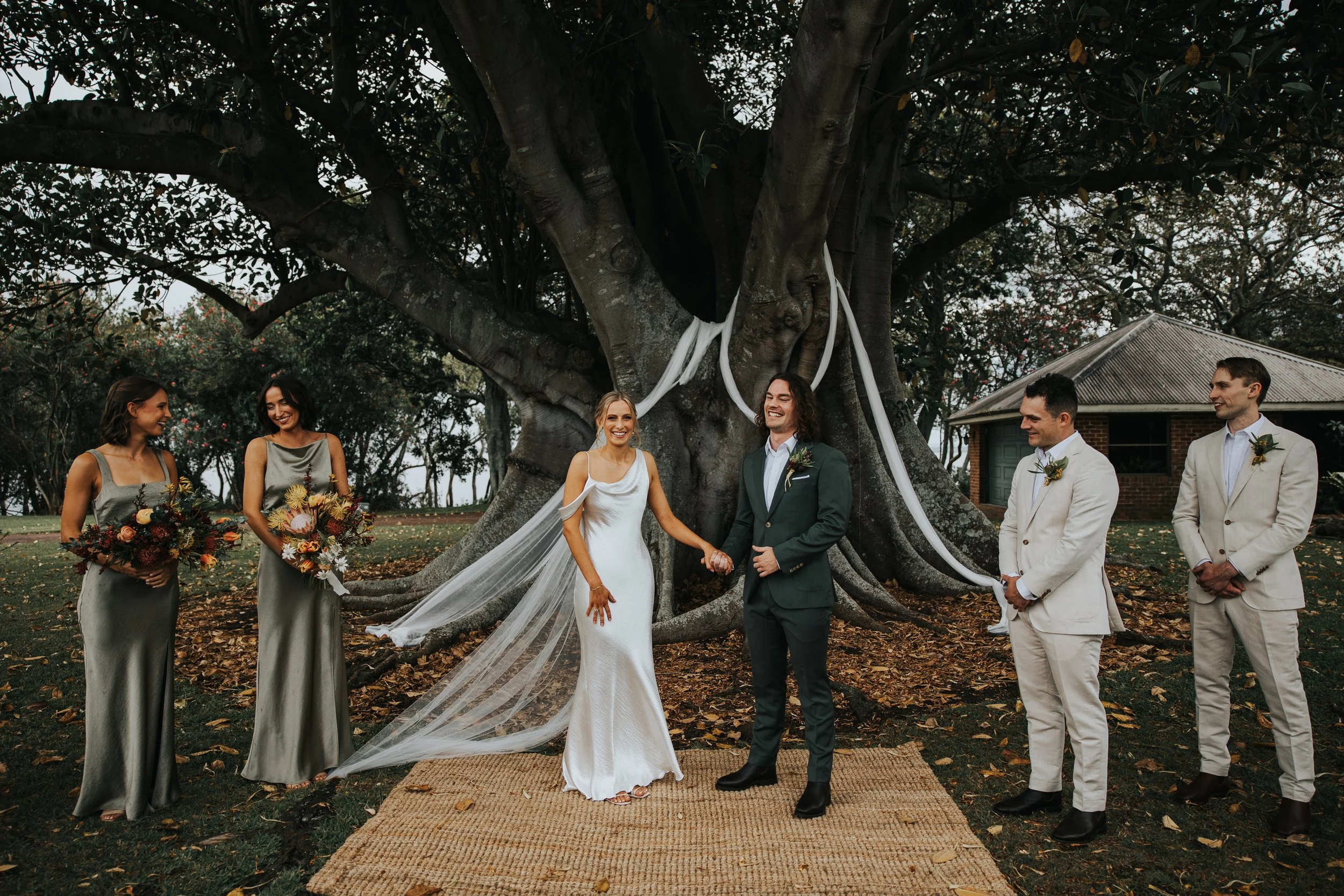 A wedding ceremony taking place outdoors under a large tree with wedding party members holding hands, including a bride and groom at the center surrounded by attendants with bouquets, a groom, and guests, with a small building in the background.