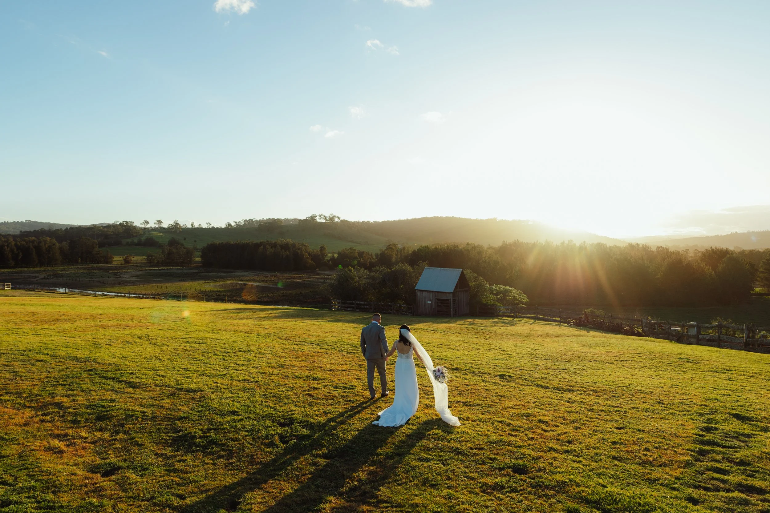 Bride and groom holding hands walking across a green field at sunset, with rolling hills and a small barn in the background.