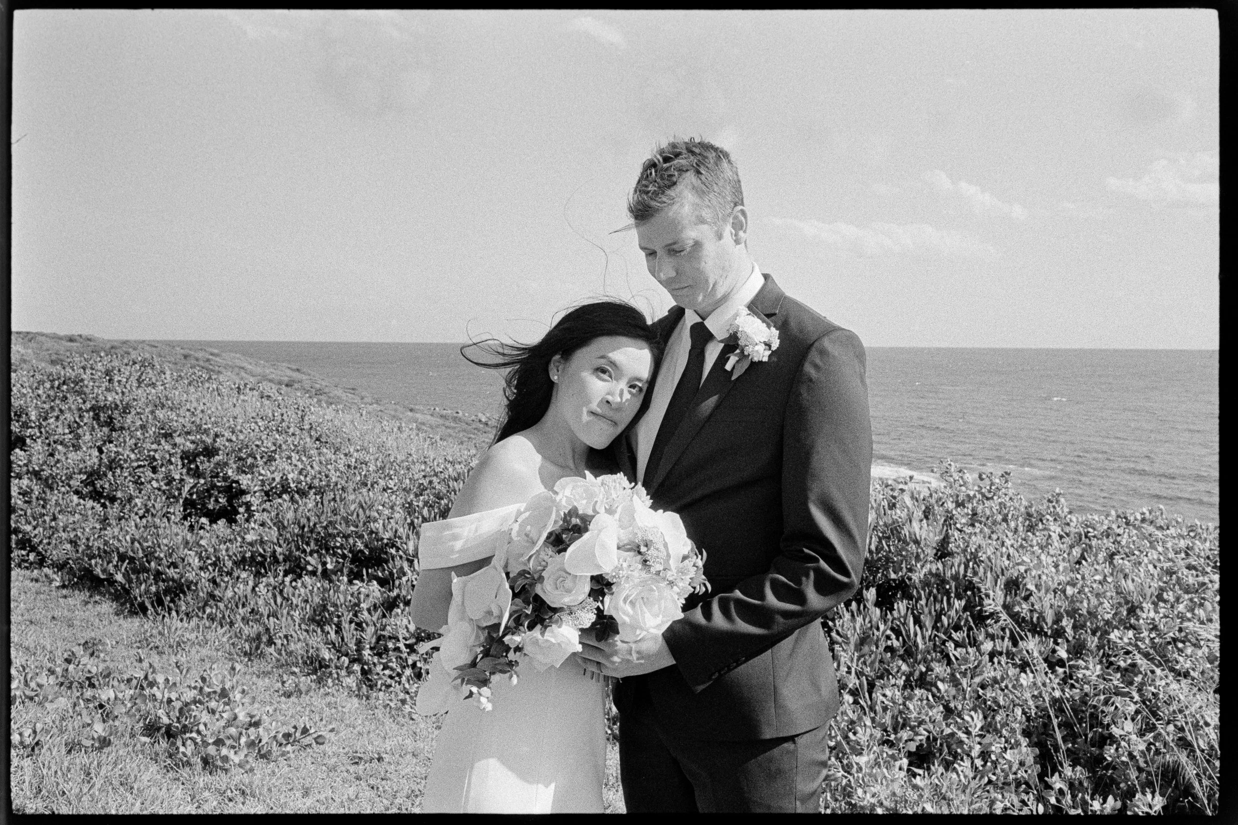 A black-and-white photograph of a bride and groom on a coastal field. The bride, holding a bouquet of flowers, leans her head on the groom's chest. The groom is dressed in a suit with a boutonniere. The ocean and cloudy sky are visible in the backgro