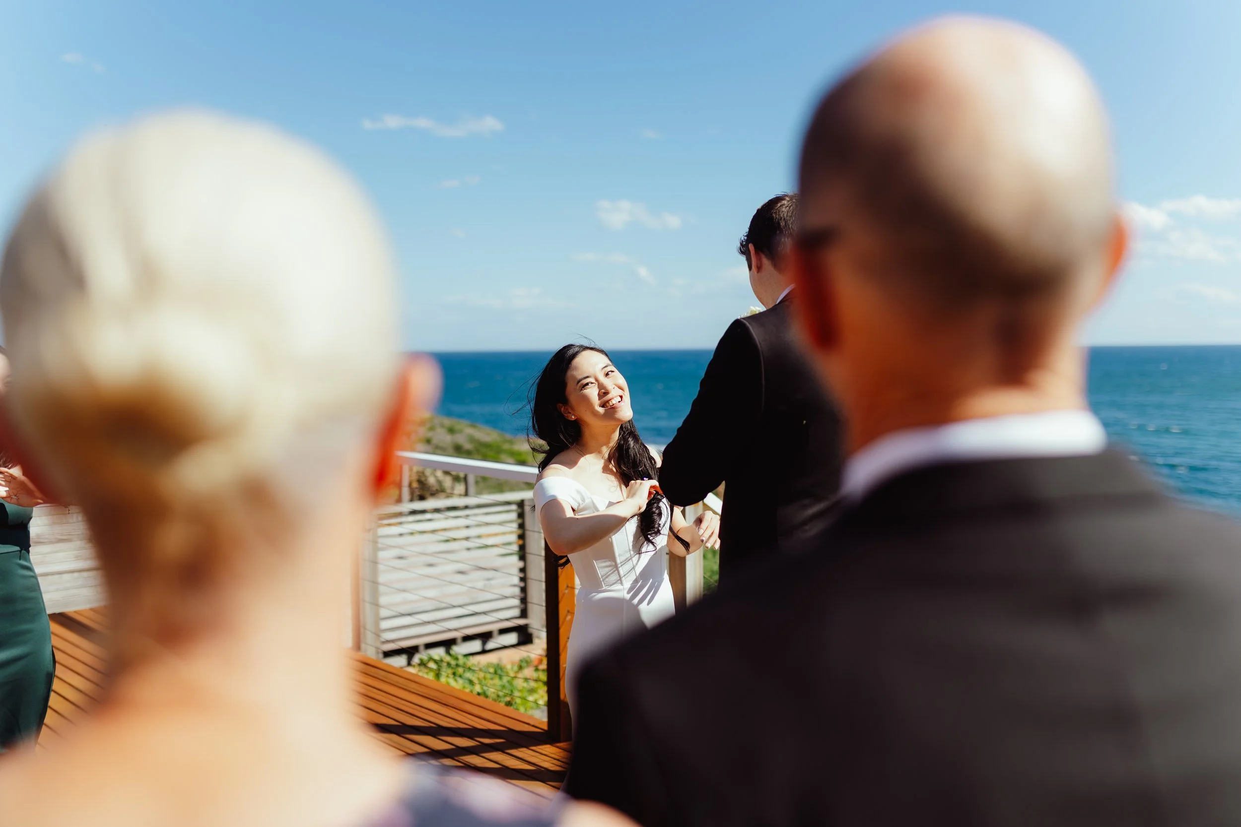 A woman in a white dress smiling at an outdoor event by the ocean with a man in a suit, while two bald men in the foreground observe.