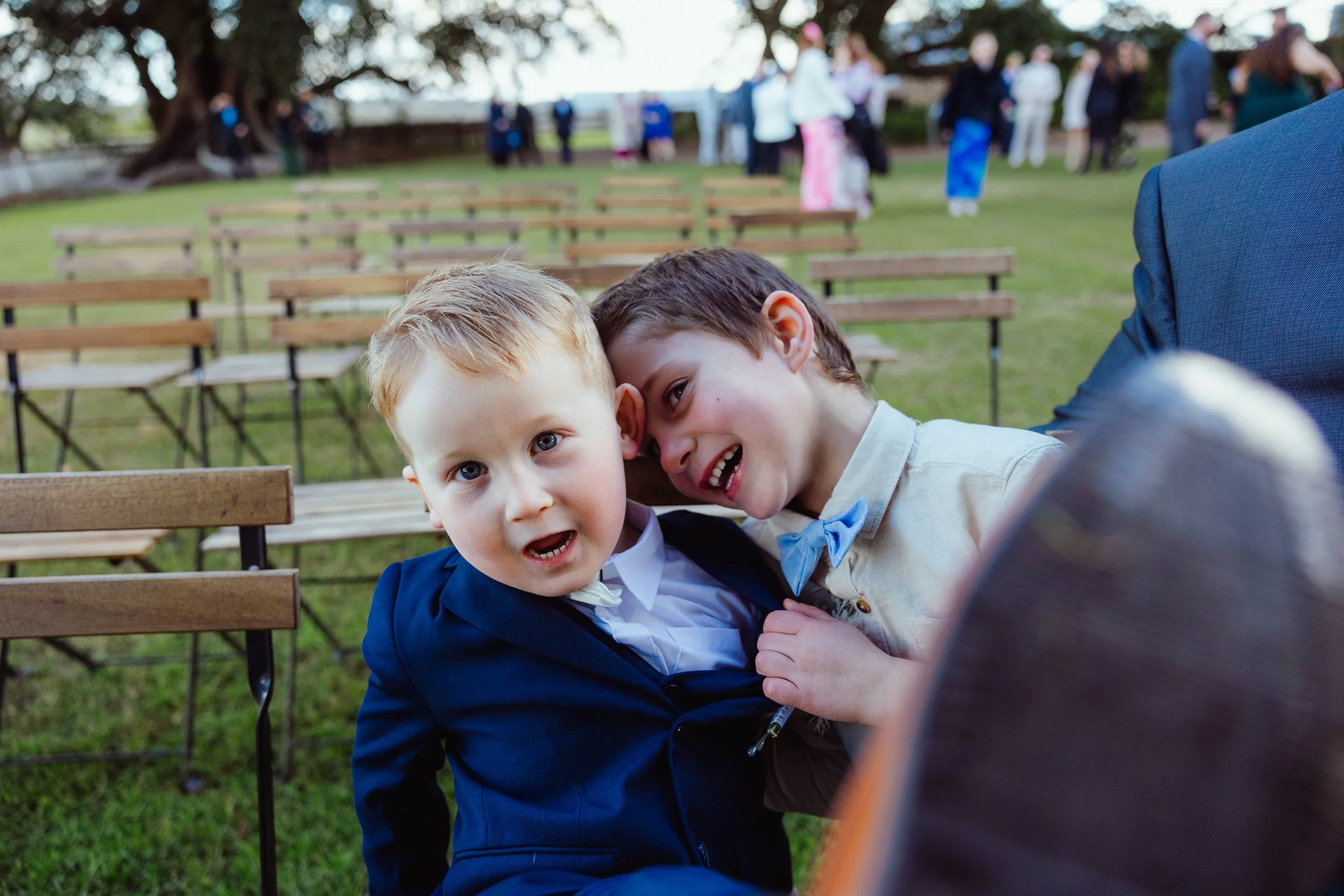 Two young boys dressed in formal attire sitting close together in outdoor setting with rows of chairs and people in the background, smiling and having fun.