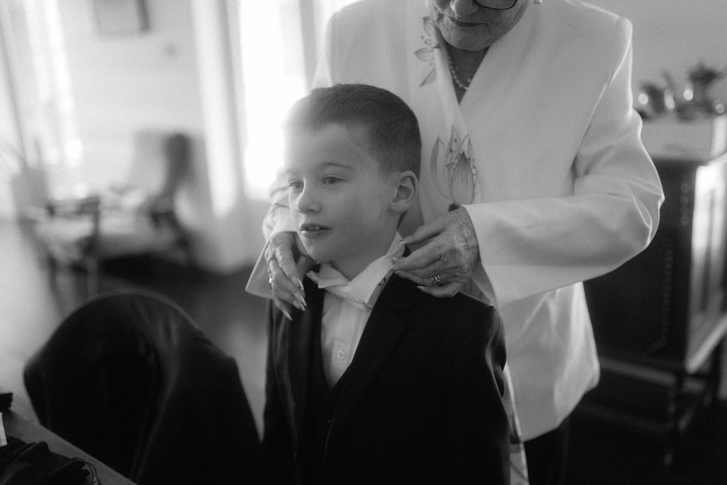 An elderly woman in a white outfit helping a young boy in a black suit with a bowtie. The woman has her hands on the boy's shoulders, and they are in a room with blurred background objects.