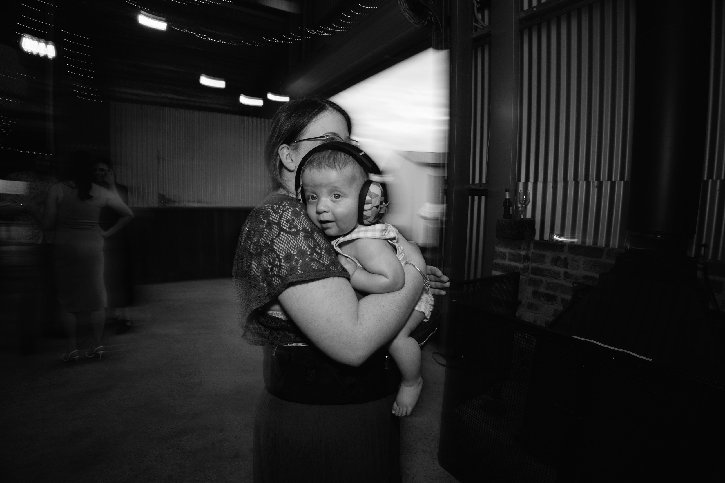 A woman holding a child with headphones at an indoor event, with other people in the background.