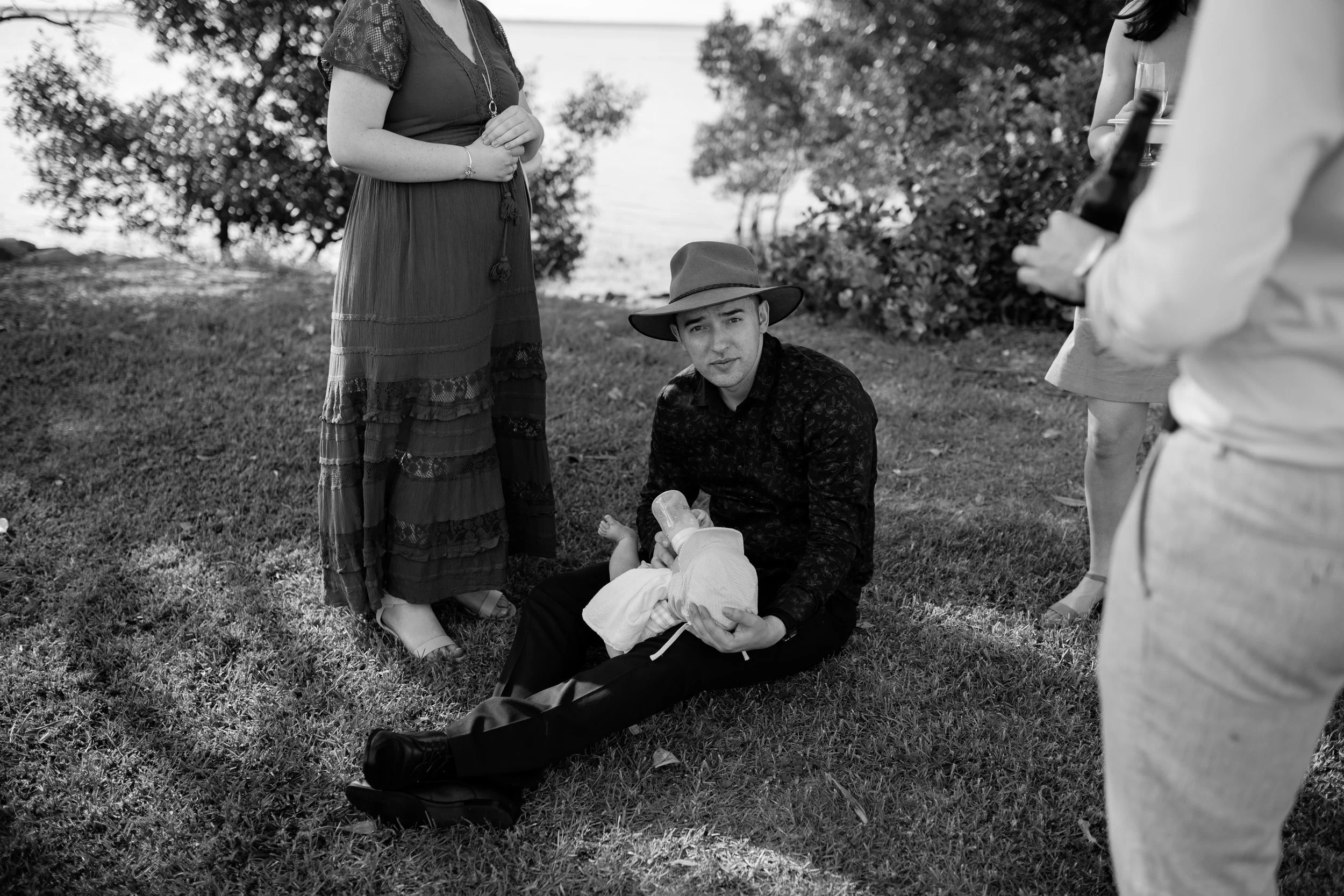 Black and white photo of a young man with a hat, kneeling on the grass, holding a baby. Several women are standing nearby, one holding a drink, with trees and a cloudy sky in the background.