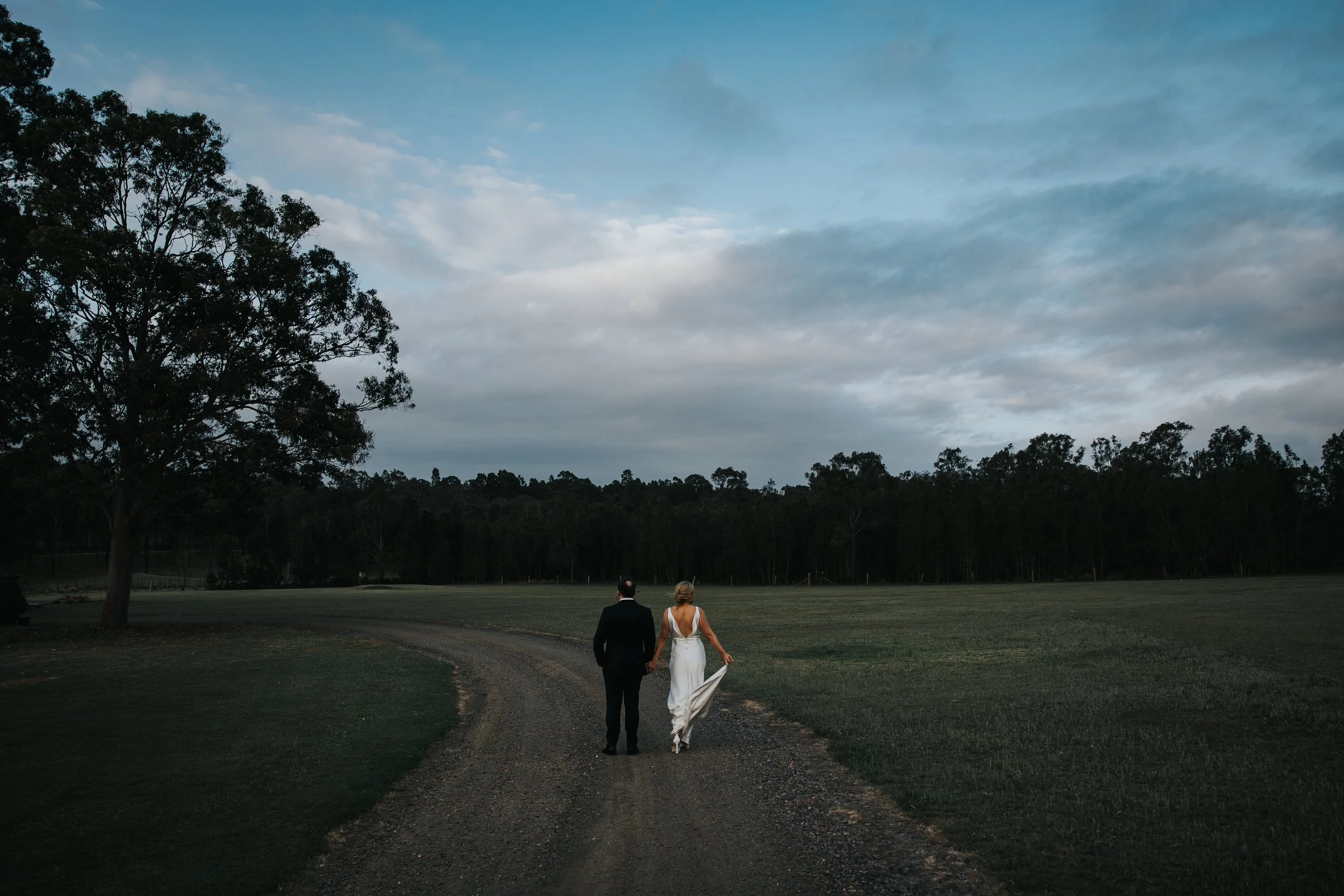 A bride and groom holding hands walk along a dirt path in a wide open field with trees and a cloudy sky in the background.
