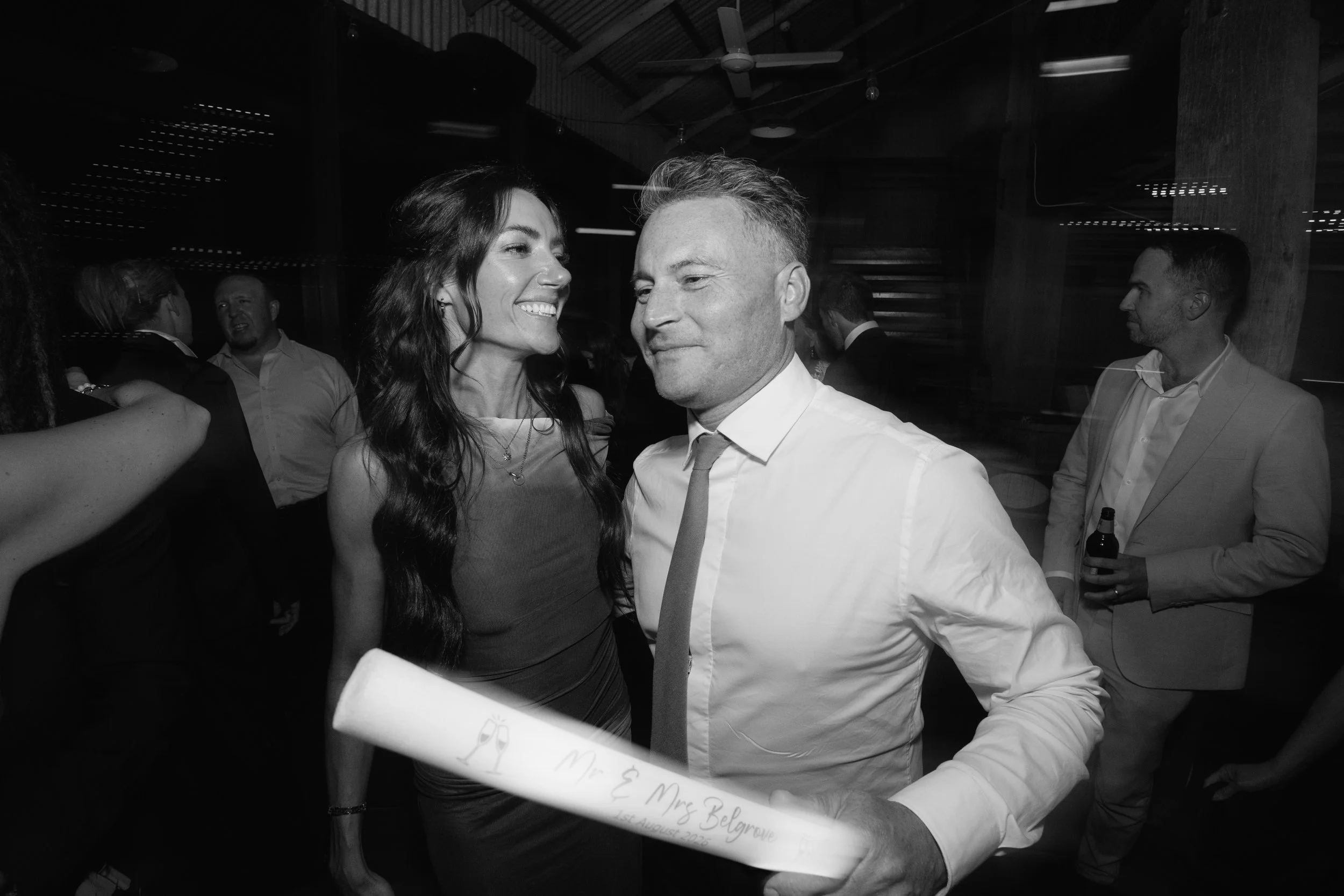 A black-and-white photo showing a couple at a wedding. The woman is smiling and facing the man, who is wearing a white shirt and tie. The scene appears to be at a reception with other guests in the background.