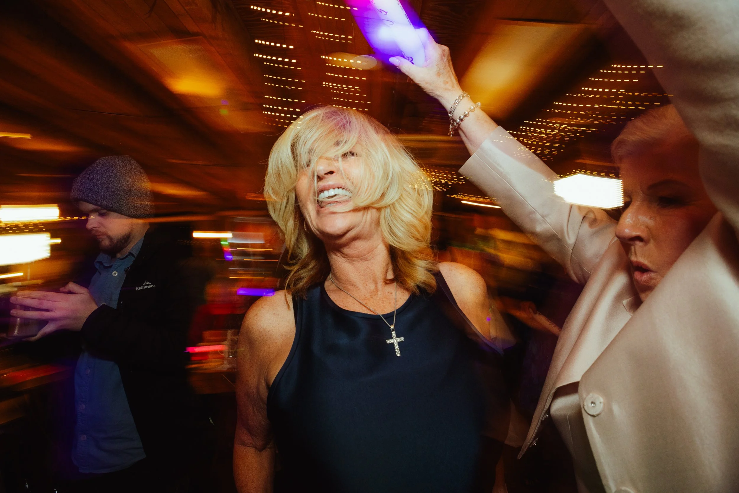 A woman with blonde hair smiling and sticking out her tongue, holding a glowing purple phone above her head at a lively party. Two other people are partially visible beside her, one with a beanie and the other with short blonde hair, in a setting wit