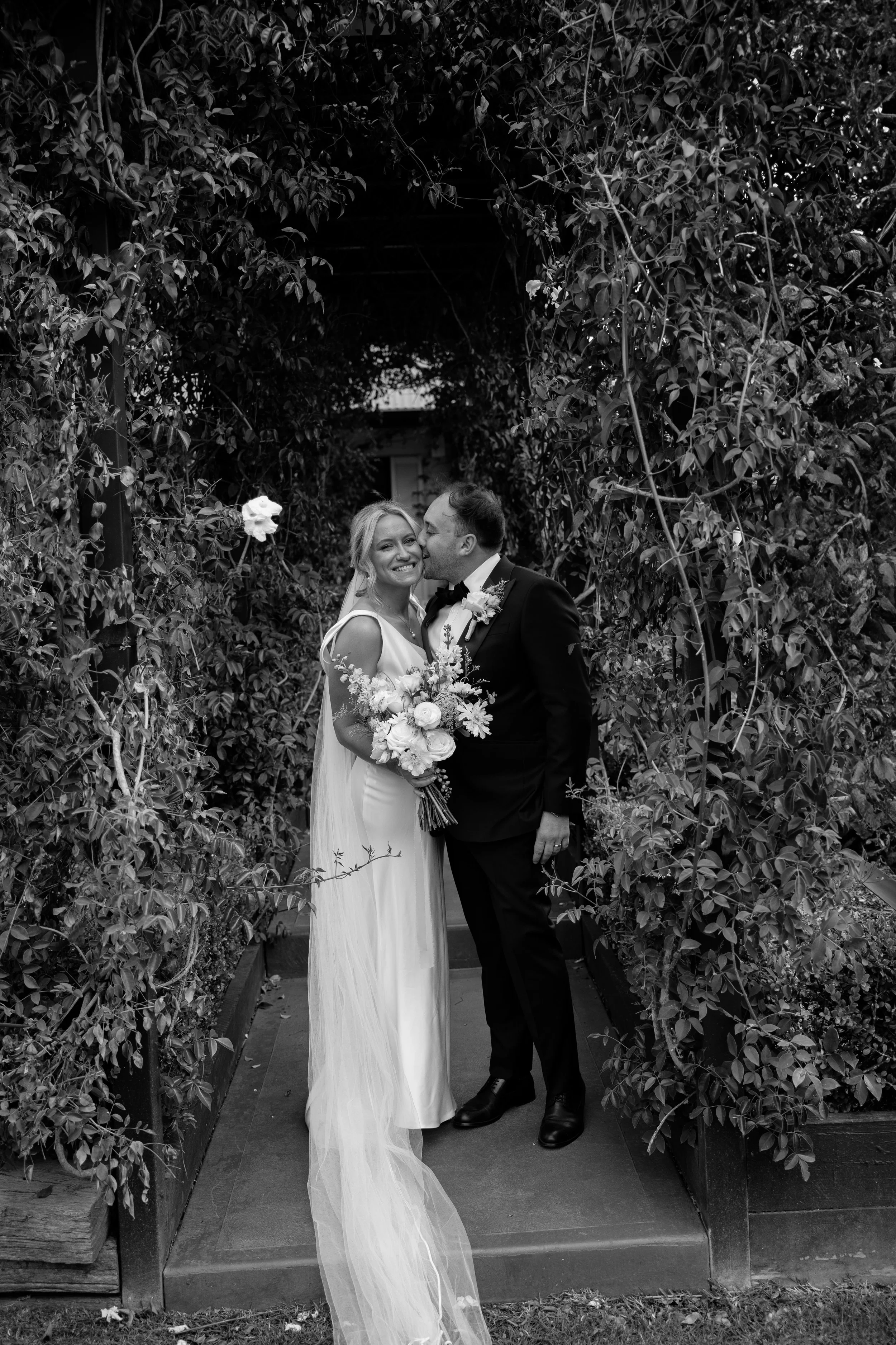 A bride and groom sharing a moment under a leafy arch, the groom kissing the bride on the cheek, both smiling. The bride is holding a bouquet and wearing a wedding gown, while the groom is in a tuxedo.