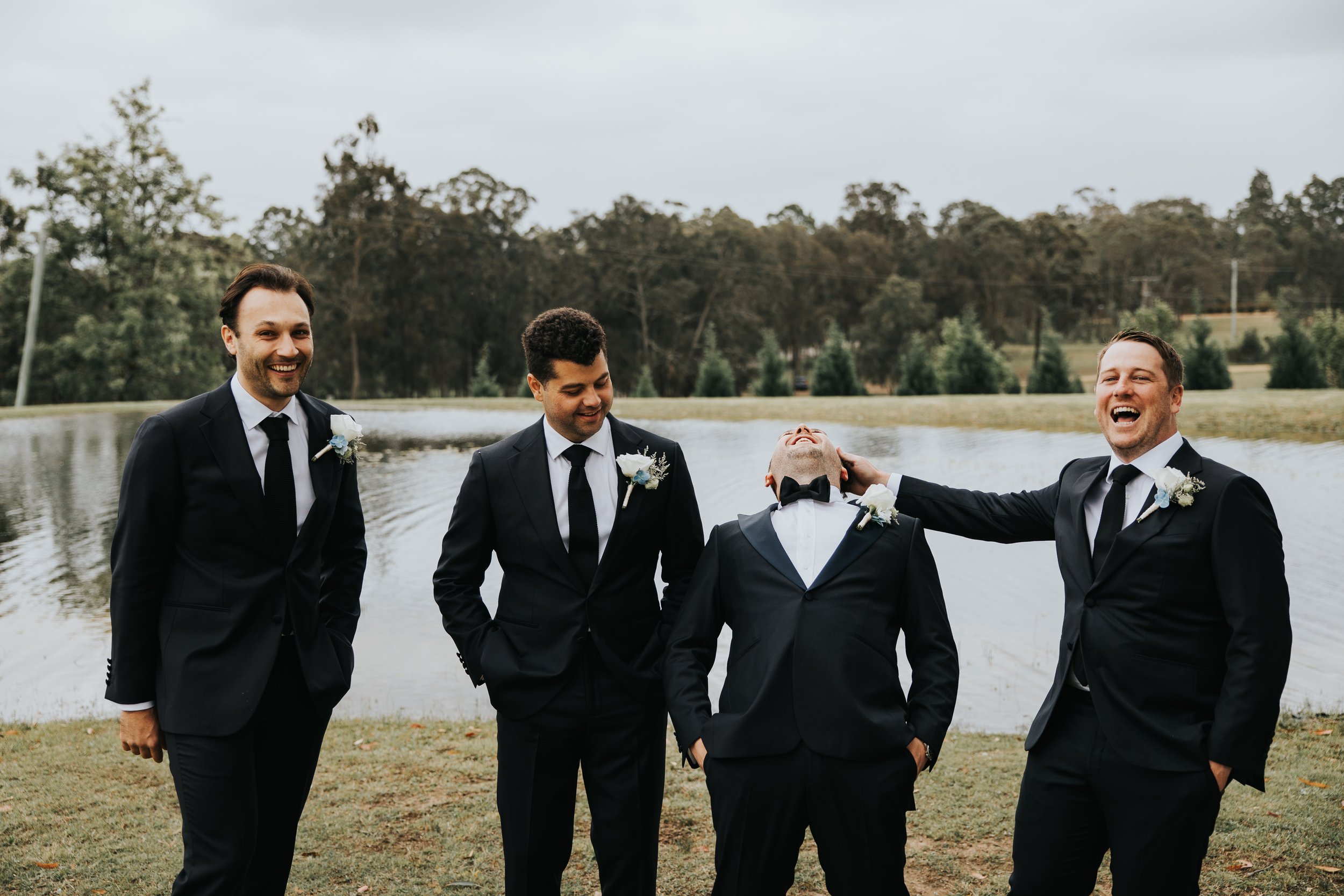 Four men dressed in black tuxedos with white shirts and black ties, standing outdoors near a lake with trees in the background, laughing and smiling.