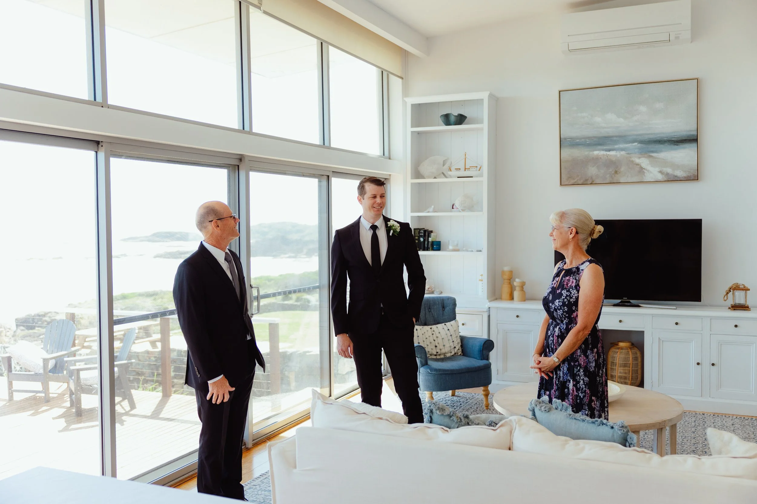 Three people, two men and one woman, are engaged in conversation inside a bright living room with large windows and ocean view. The man on the left is bald and wearing glasses, the man in the middle is young with brown hair in a black suit with a whi