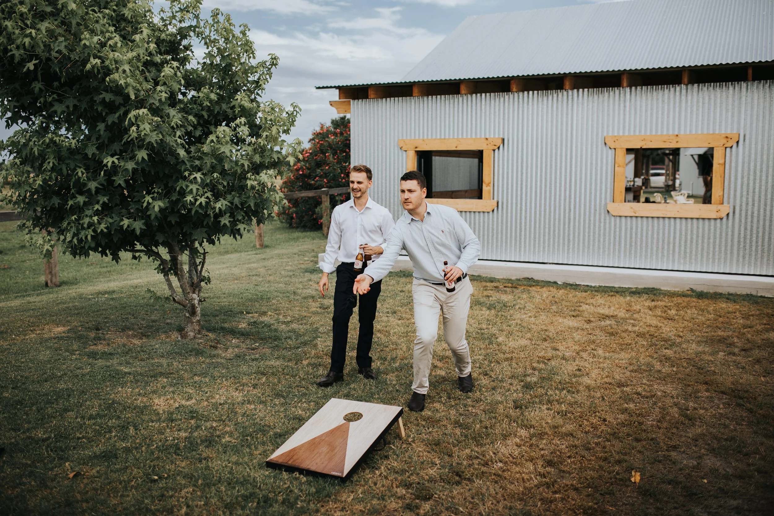 Two men playing cornhole outside near a metal building with wooden window frames and a tree.
