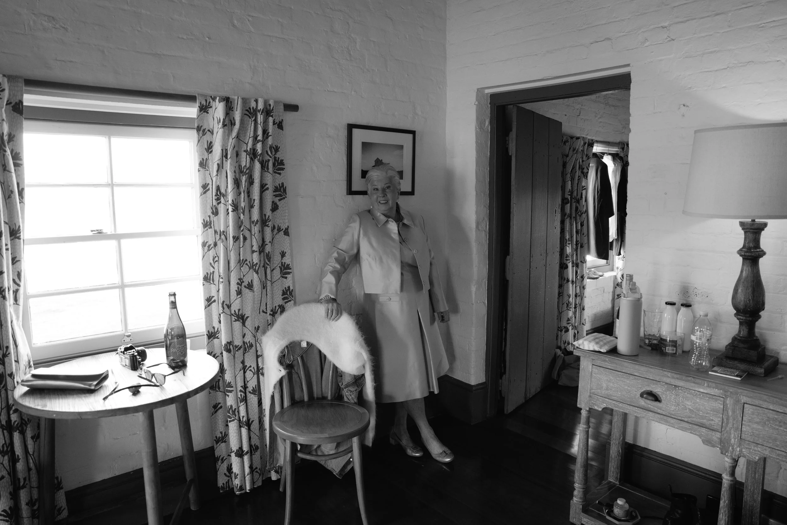 Black and white photo of an elderly woman standing in a cozy, rustic room with white brick walls. She is smiling, dressed in a light-colored coat, and is standing near a dining table with a bottle, glasses, and papers. The room has floral curtains, a