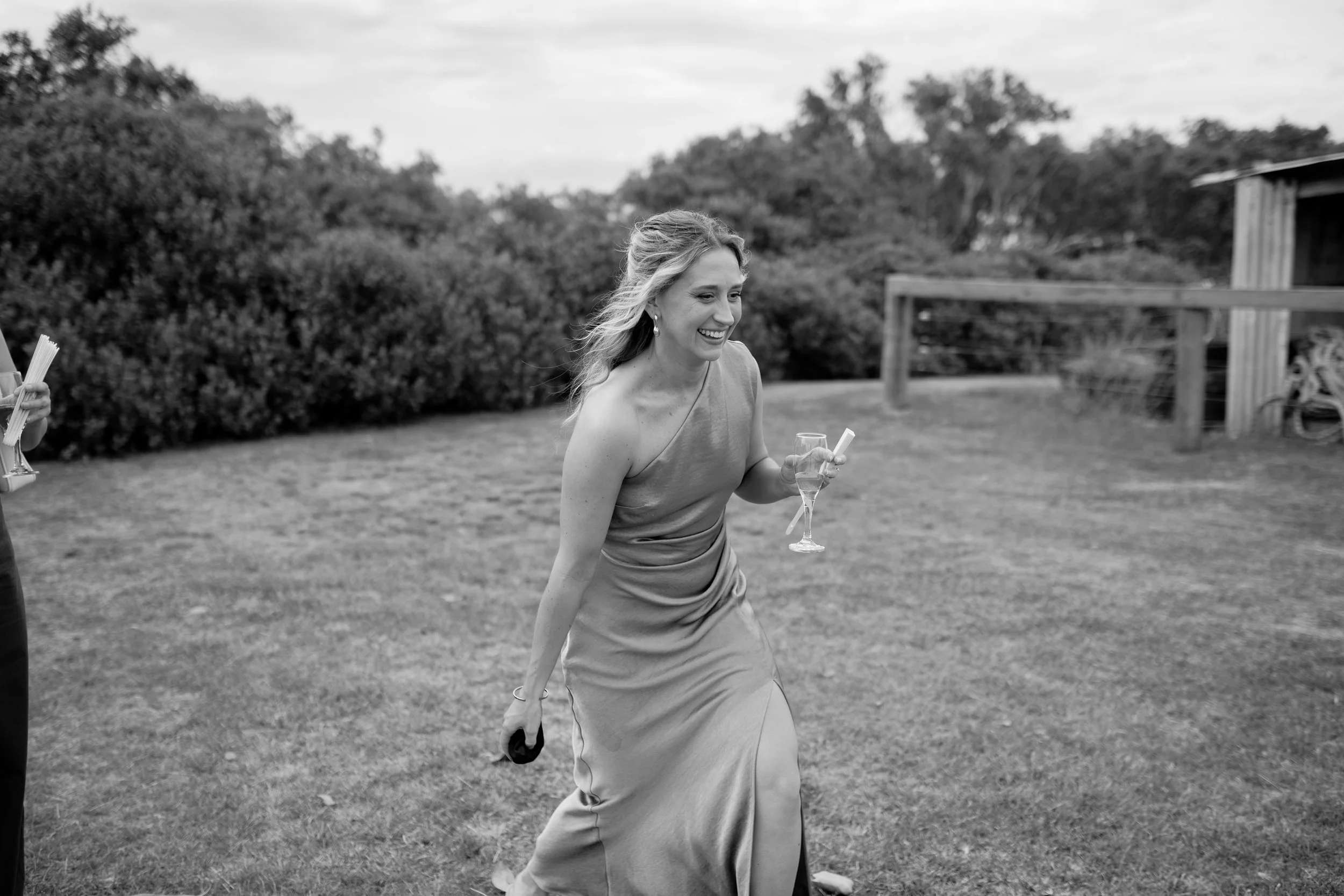 A woman in a strapless dress holding a glass of champagne and a small gift box, smiling and walking outdoors on grass with trees and a wooden structure in the background.