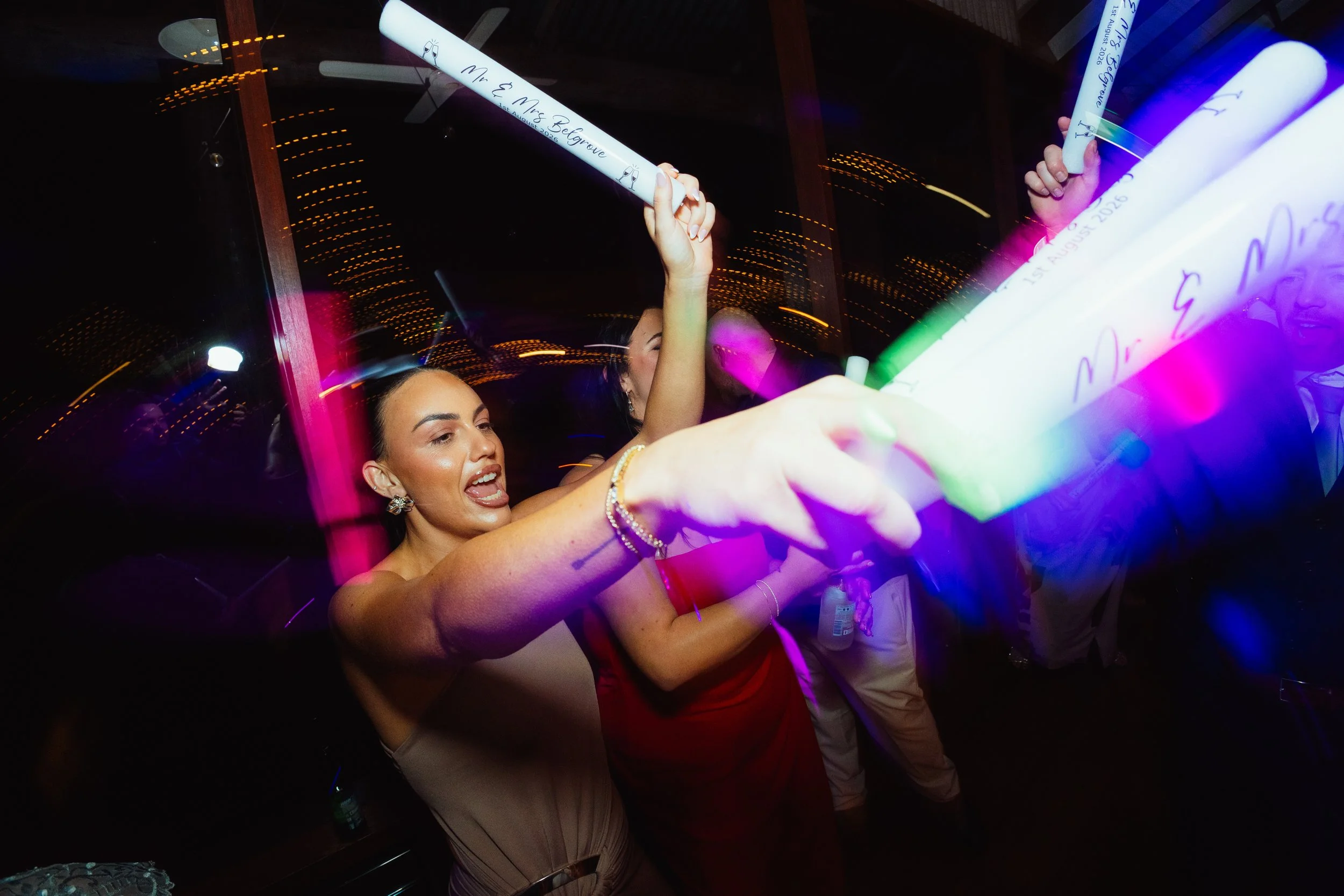 People celebrating at a wedding reception, holding illuminated glow sticks with names and dates, dancing and enjoying the event in a dimly lit venue with reflections on glass windows.