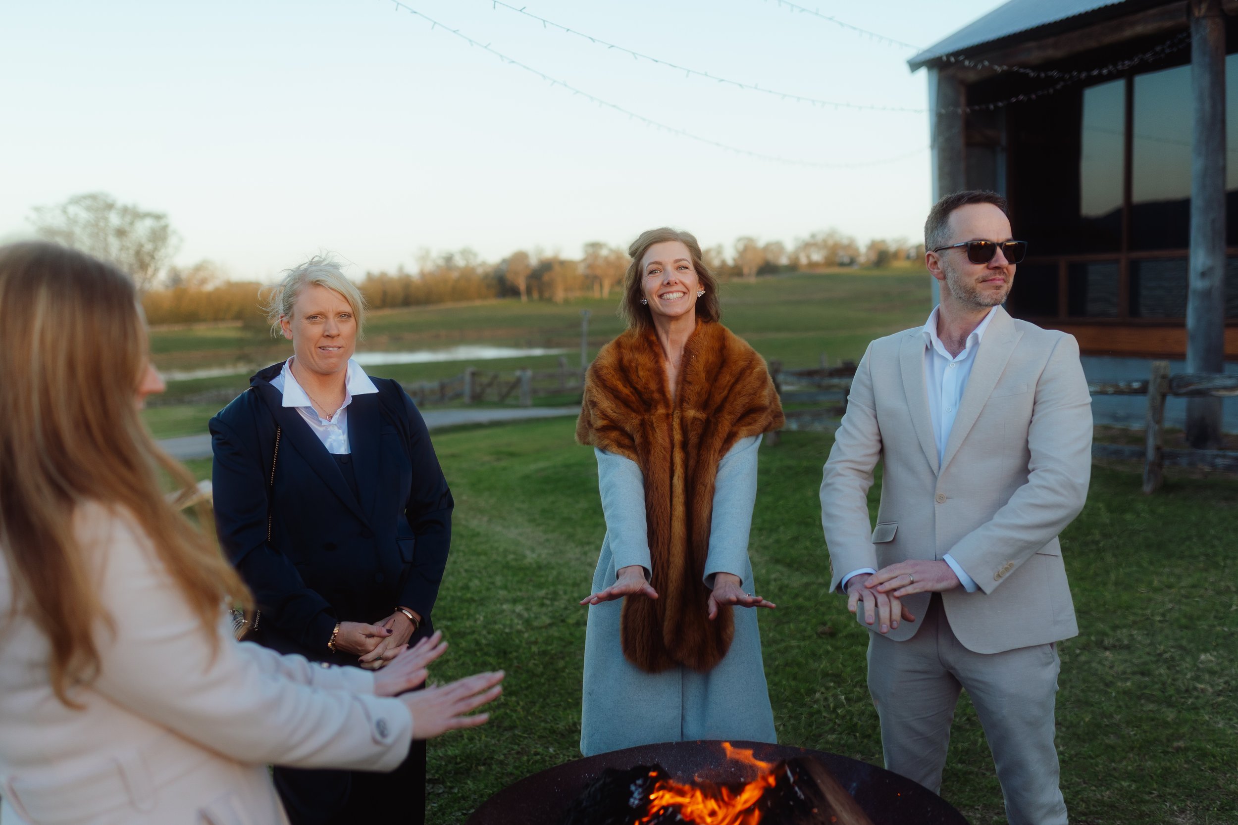 People gathered around a small outdoor fire pit, chatting on a grassy area with trees and a fence in the background, during sunset.