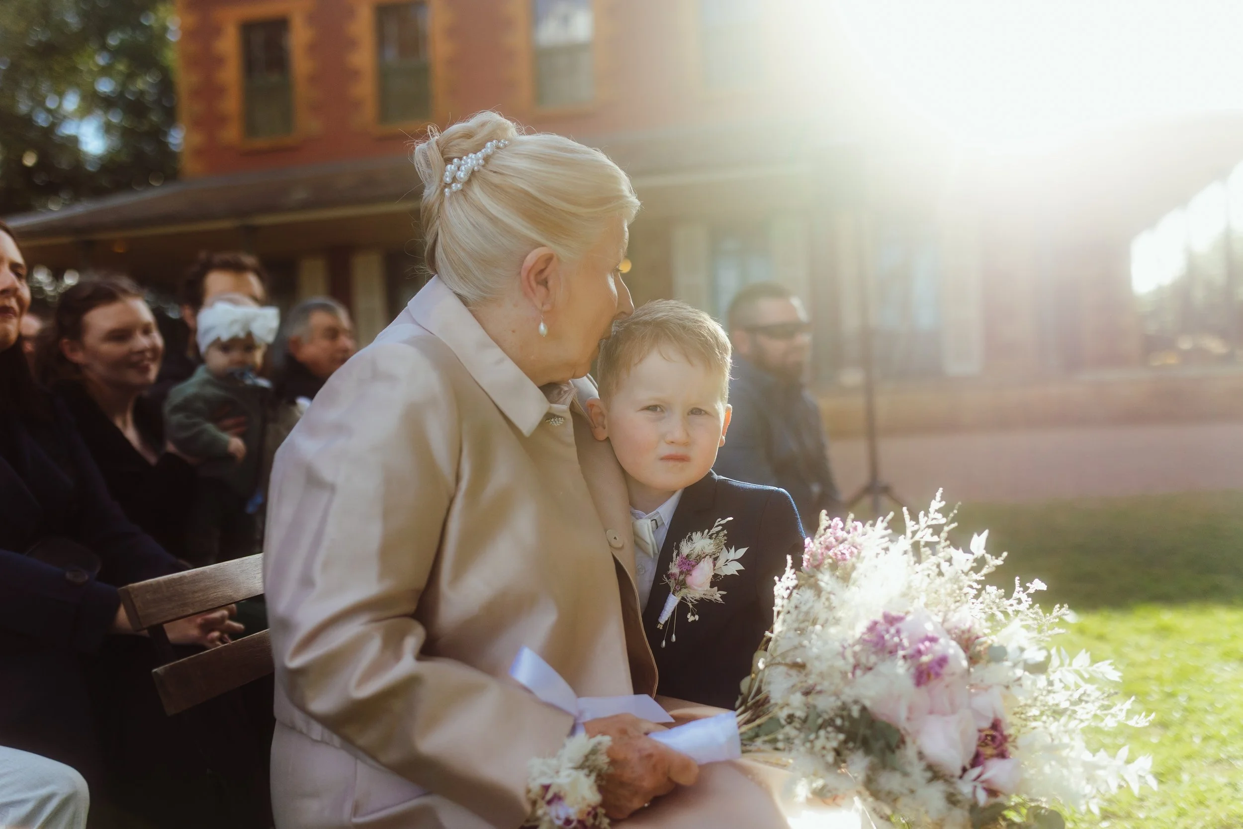 An elderly woman with a pearl hair accessory and pearl earrings, sitting outdoors with a young boy, holding a bouquet of white and pink flowers. Several people are in the background, some smiling, in a sunny setting with a brick building in the dista