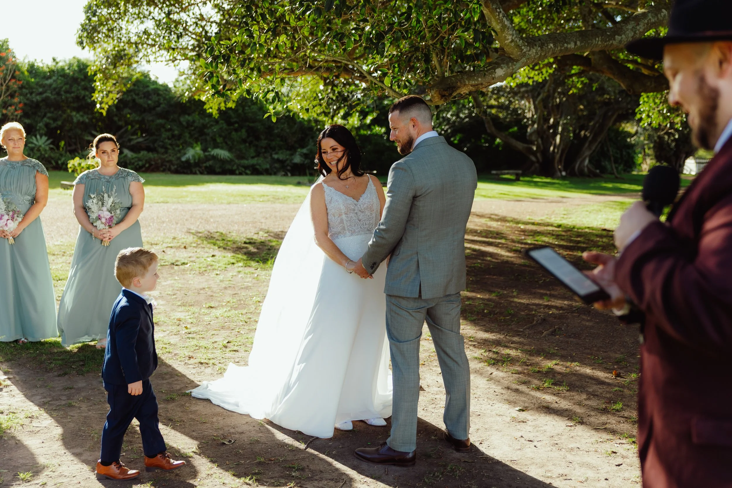 A wedding ceremony outdoors with a bride and groom holding hands, surrounded by bridesmaids and groomsmen, under a large tree on a sunny day.