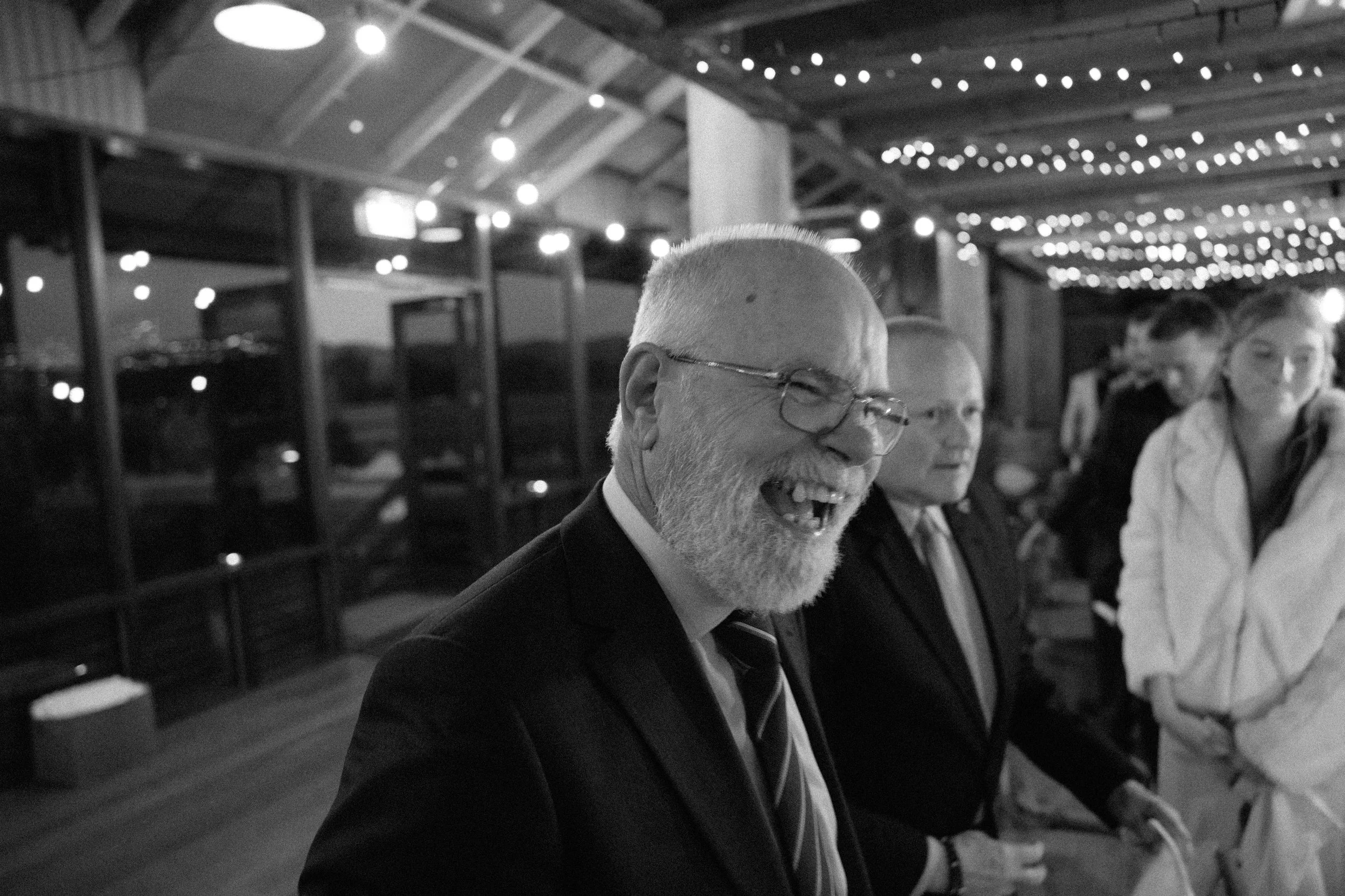 A black-and-white photo of an older man with glasses and a beard, smiling and laughing, at a social gathering. There are other people in the background, and the ceiling is decorated with string lights.