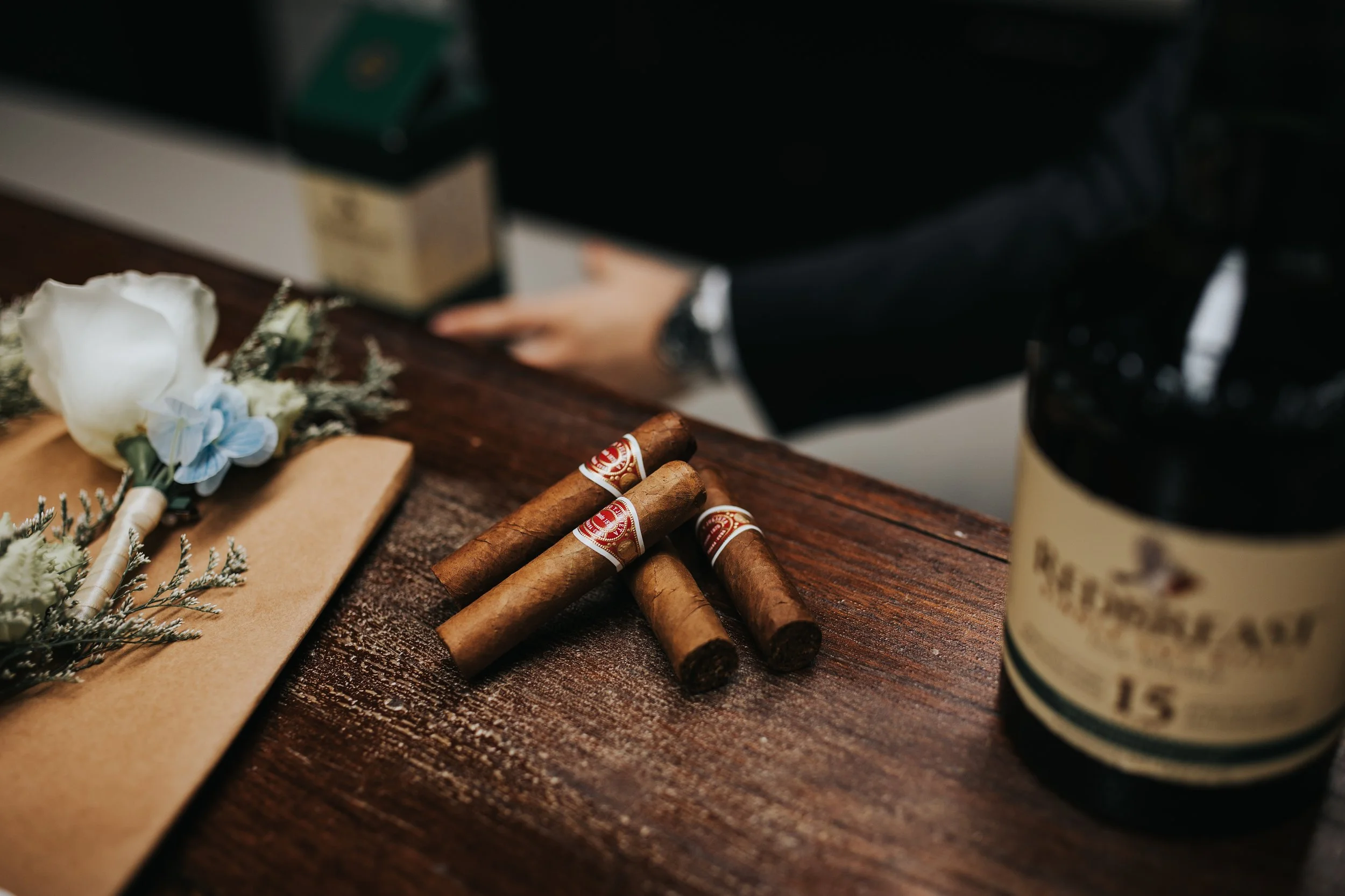 Four cigars with red and gold bands on a wooden table, with a blurred black coffee mug and some flowers in the background.