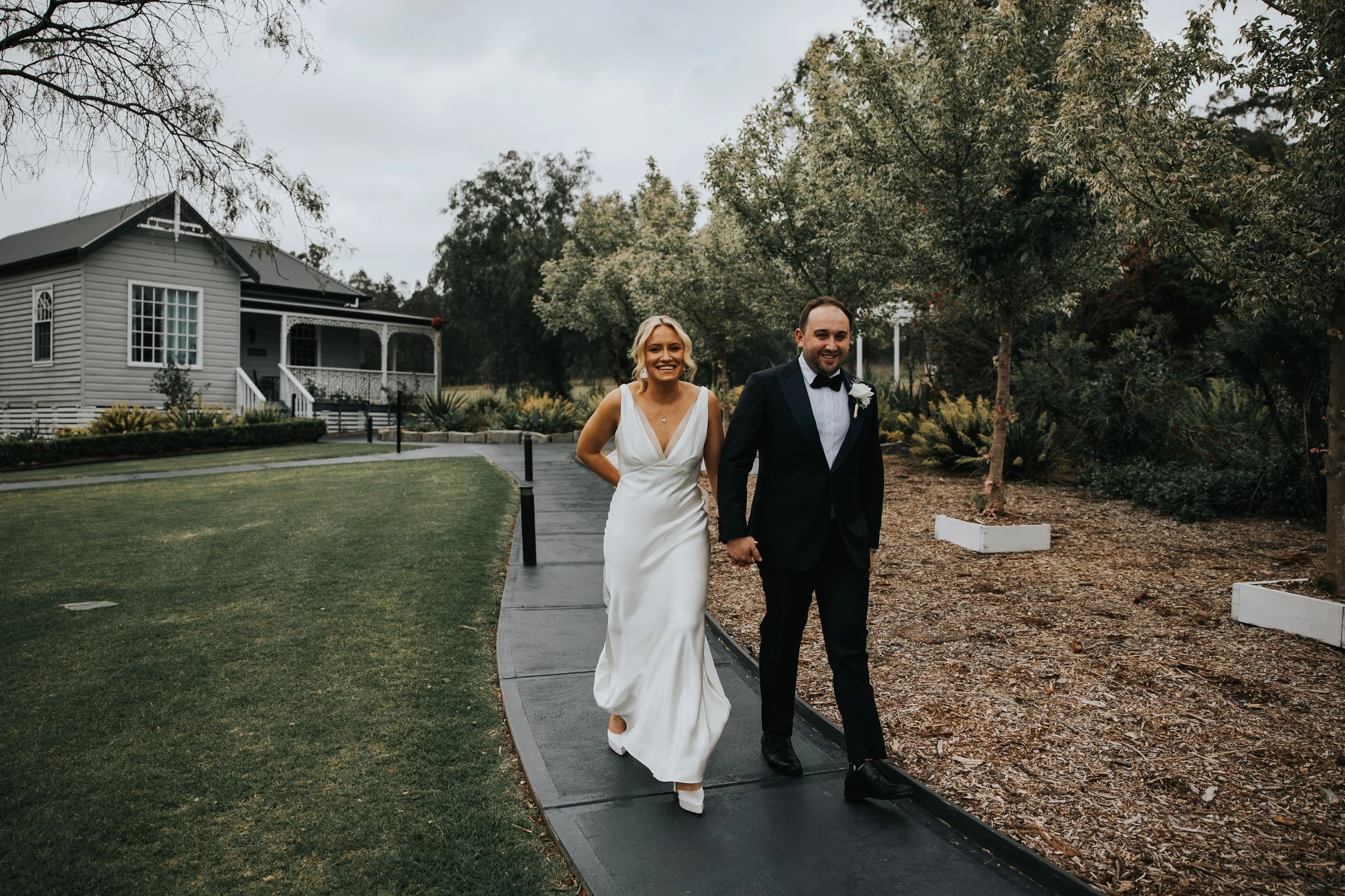 A bride and groom walking hand in hand on a paved pathway outside, with trees and a house in the background. The bride is wearing a white wedding gown and the groom is in a dark tuxedo. They are smiling.