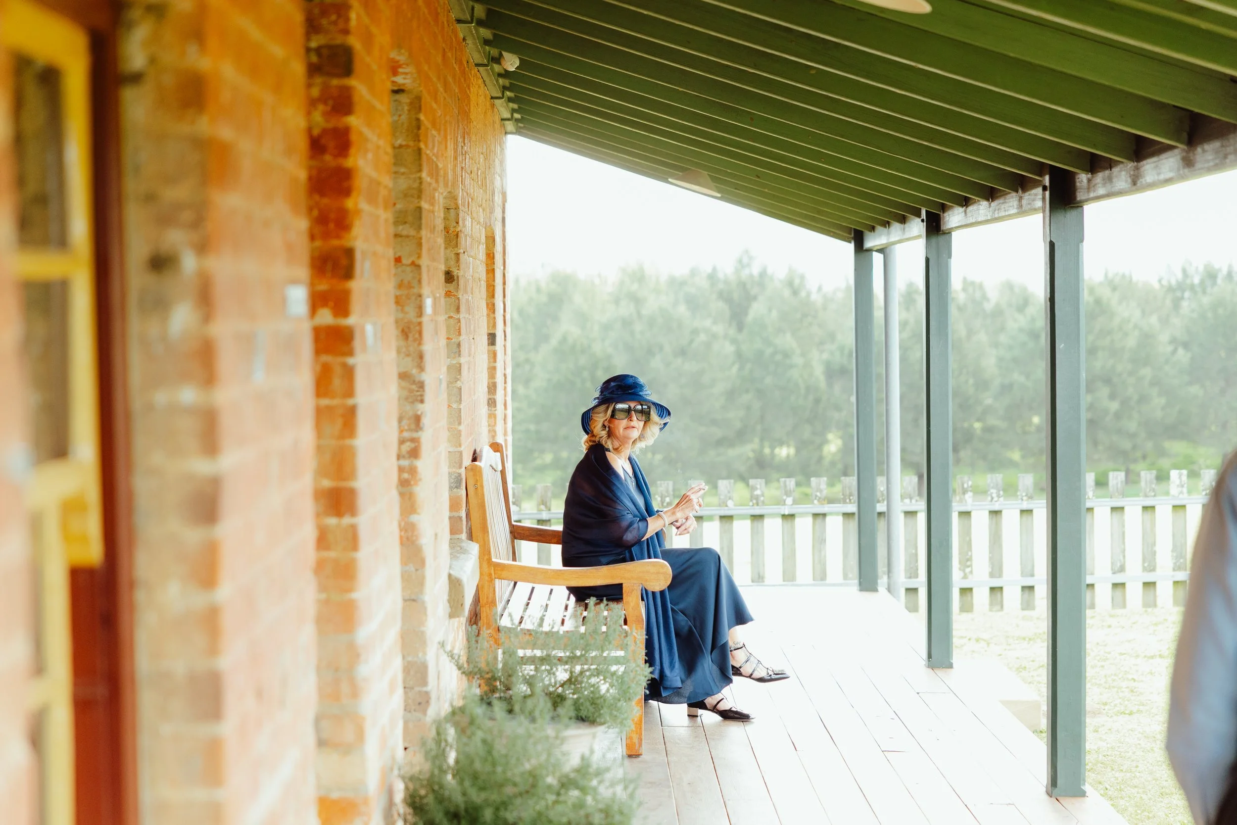 A woman in a blue dress and wide-brimmed hat, wearing sunglasses, is sitting on a porch with wooden chairs beside a brick wall, with a view of trees and a sunny sky in the background.