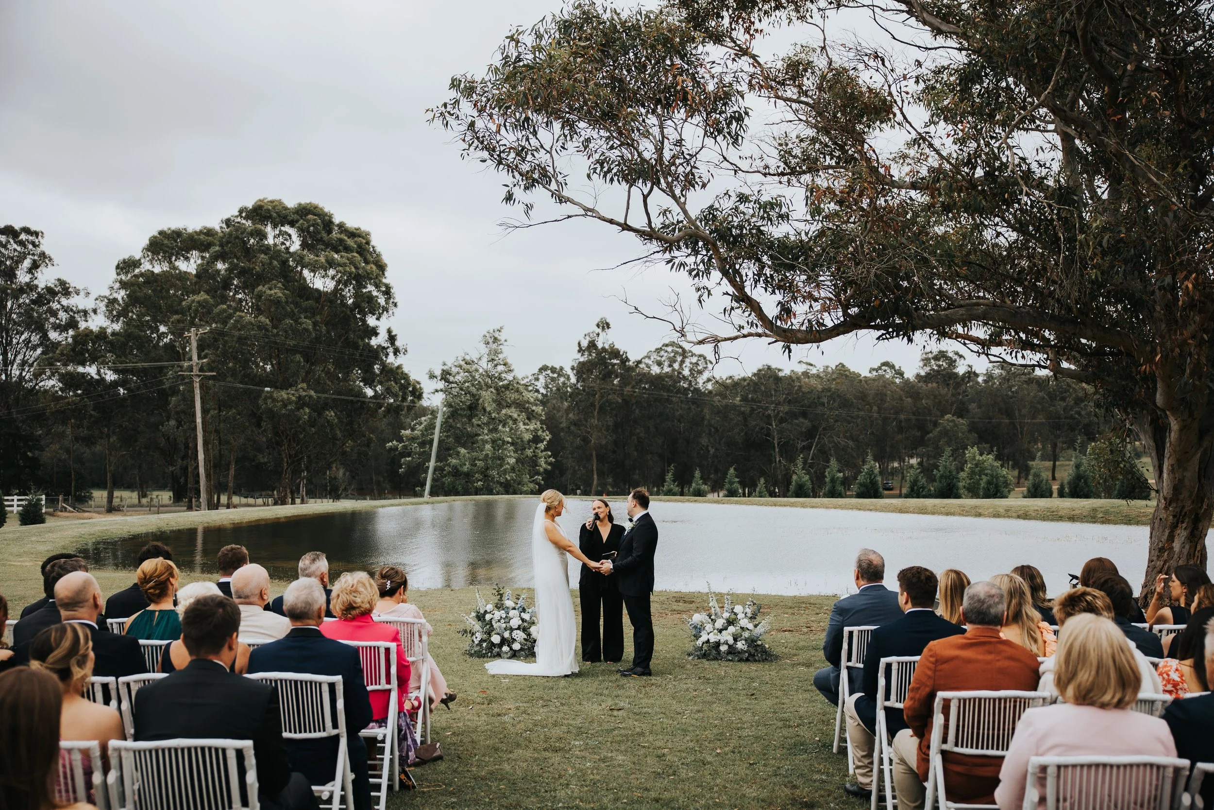 A wedding ceremony taking place outdoors by a pond, with a bride and groom holding hands in front of an officiant, surrounded by seated guests, trees, and an overcast sky.