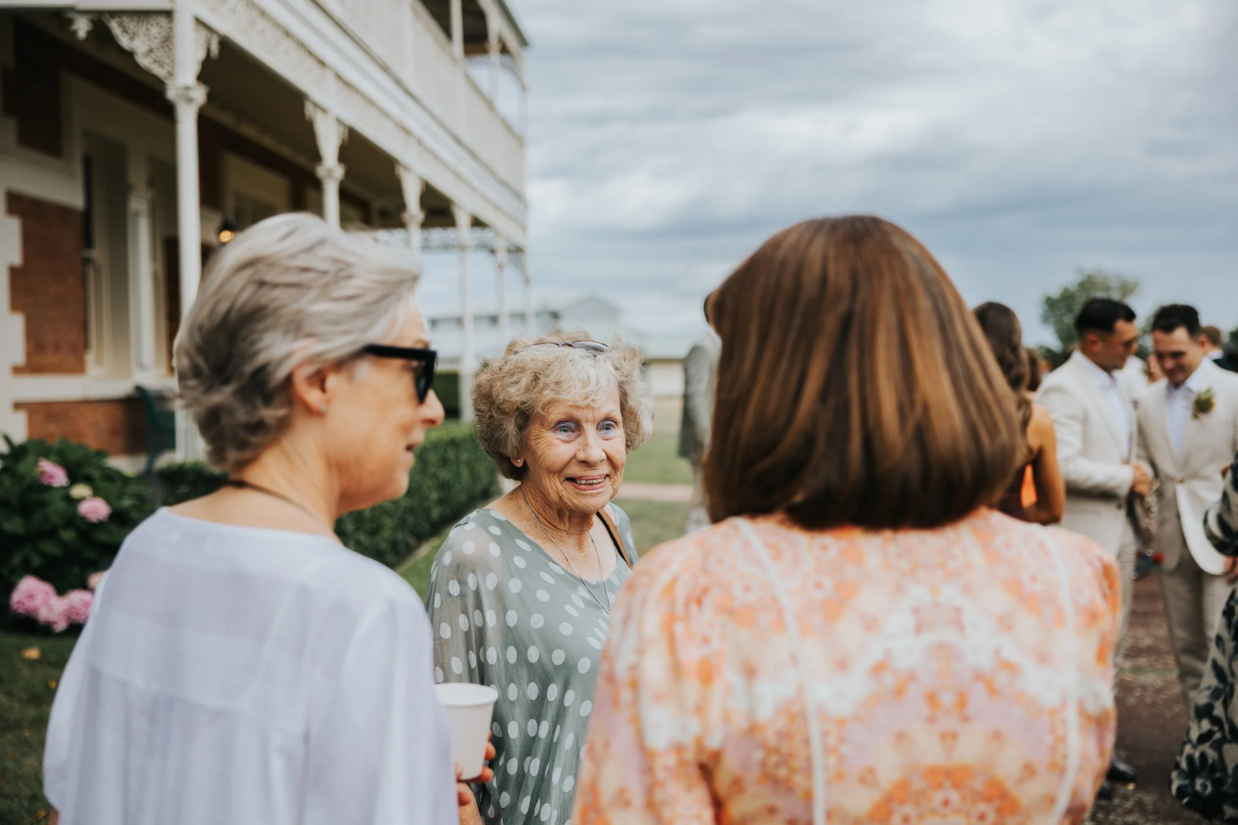 A group of women socializing outdoors at a gathering, with a large house in the background. The women are engaged in conversation.