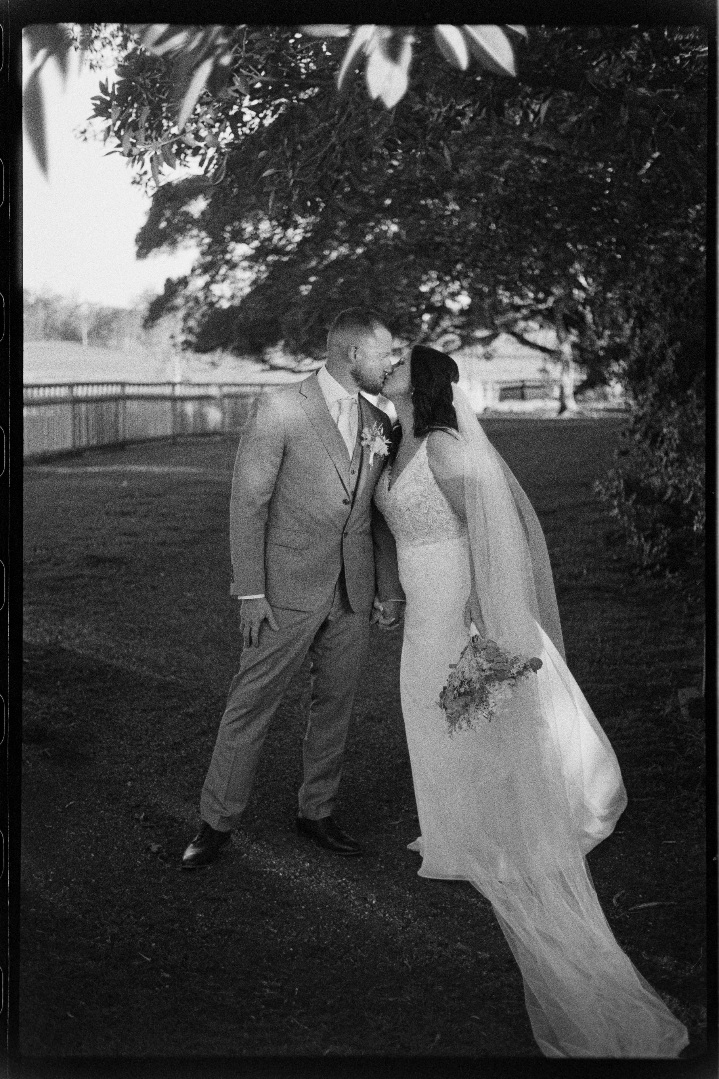 A black and white photo of a newlywed couple kissing under a large, leafy tree outdoors, with the bride holding a bouquet and wearing a long gown, and the groom in a suit.