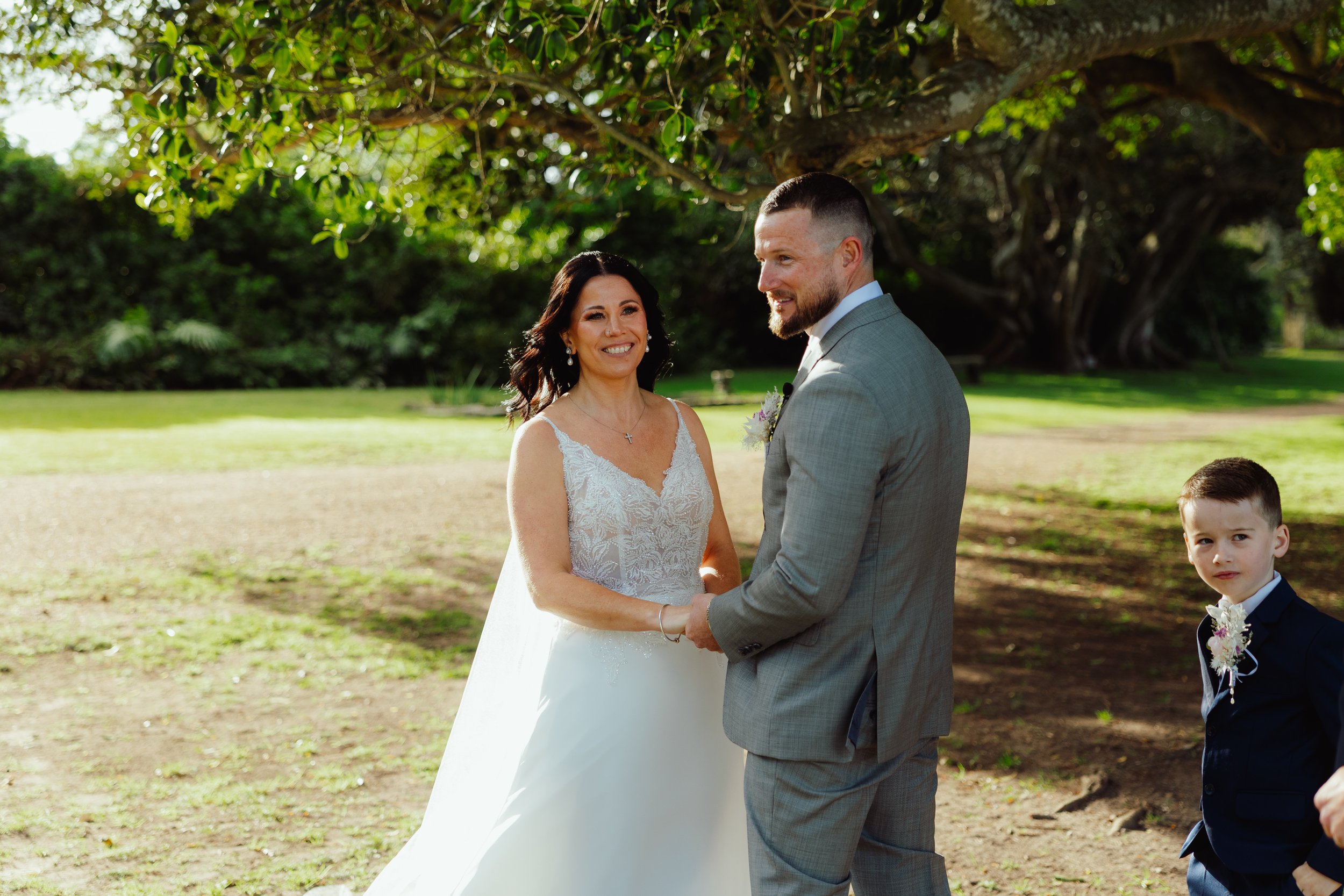 A bride and groom standing outdoors under a tree, holding hands during a wedding ceremony, with a young boy dressed in a dark suit and white shirt standing nearby.
