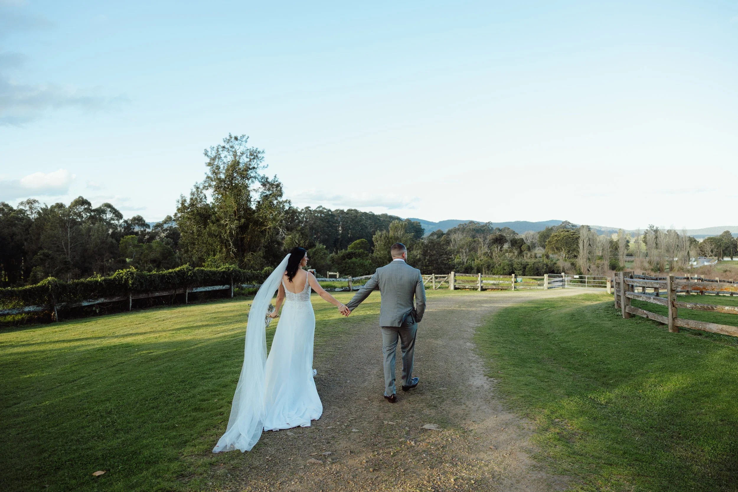 A bride and groom walking hand in hand on a grassy path outdoors, with trees and distant hills in the background, during daylight.
