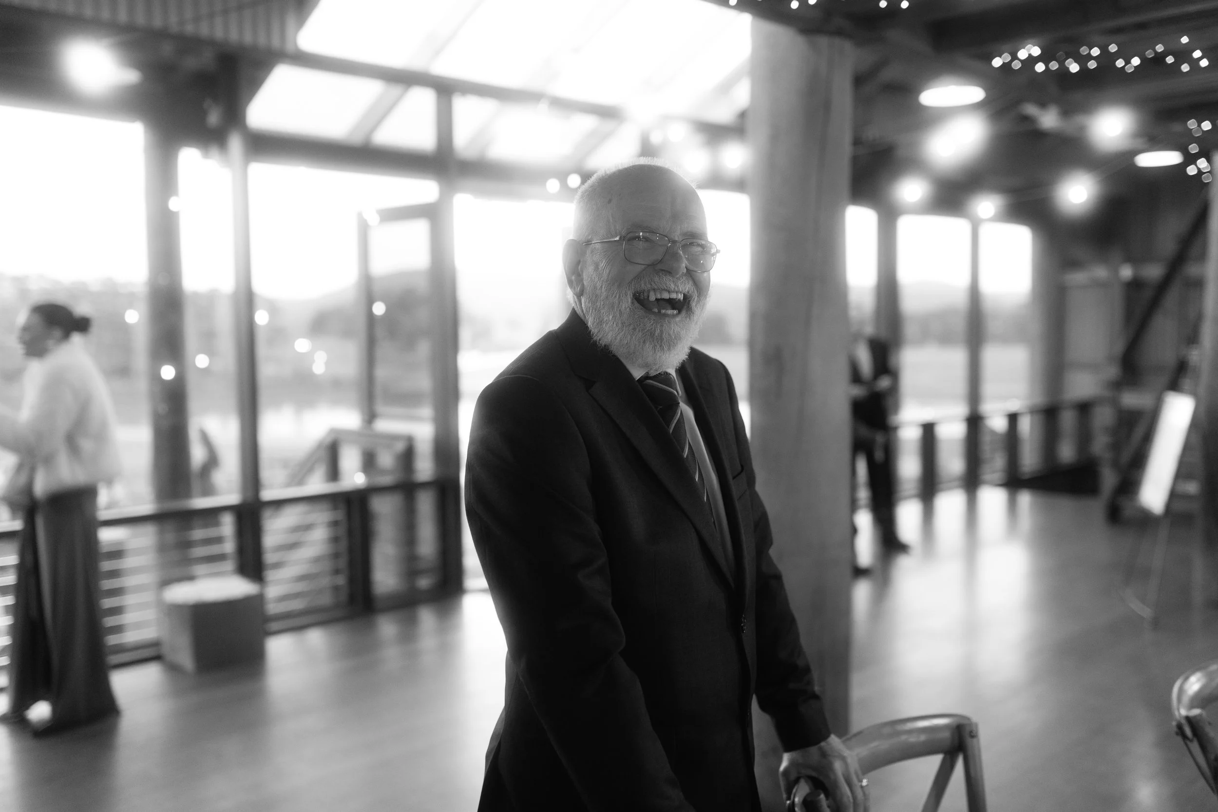 A cheerful elderly man with glasses and a beard, wearing a suit, standing inside an airport terminal. He is smiling and appears happy. In the background, a woman wearing a white blazer is visible.