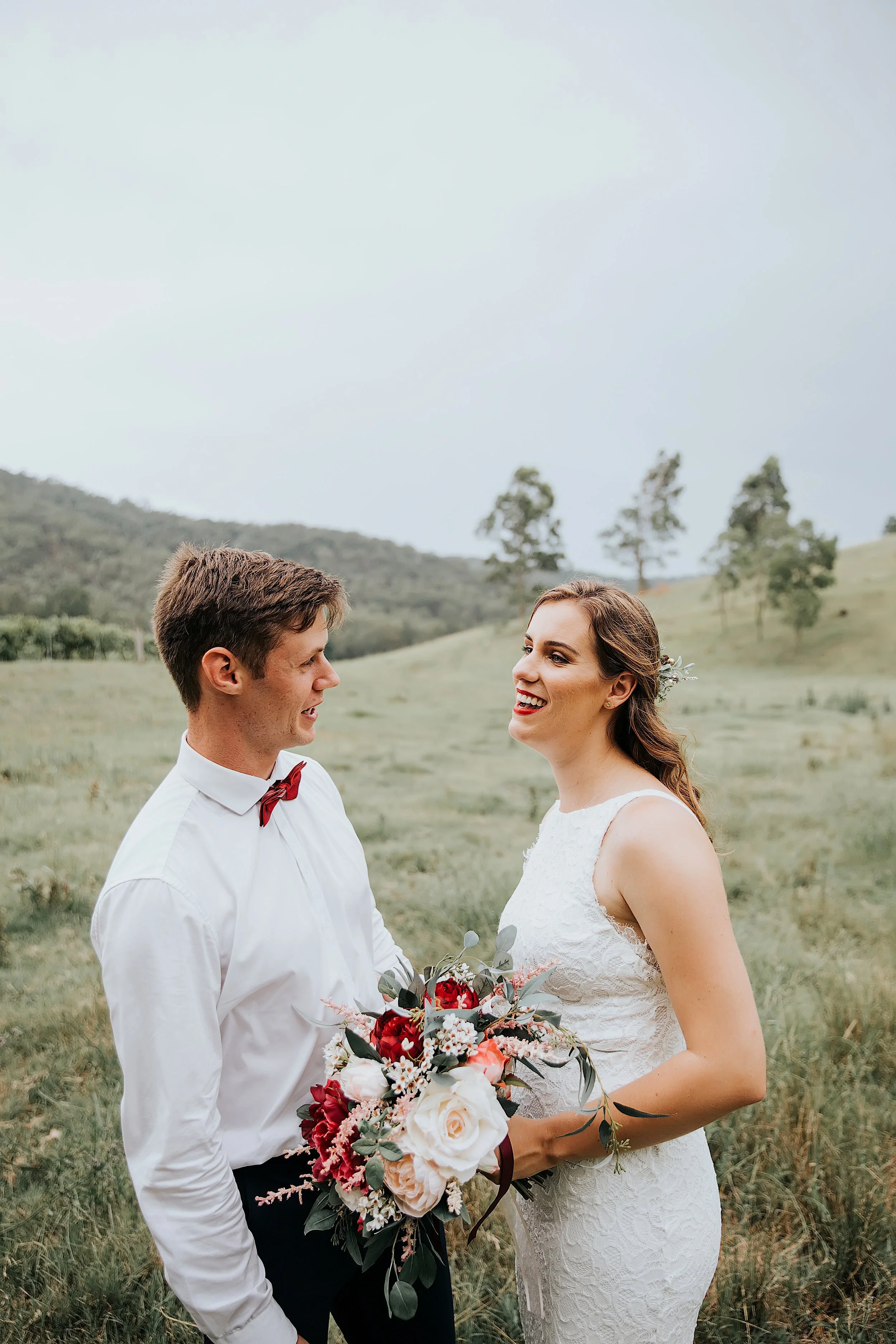 A bride and groom standing in a grassy field with hills and trees in the background, holding a bouquet of flowers, and smiling at each other.