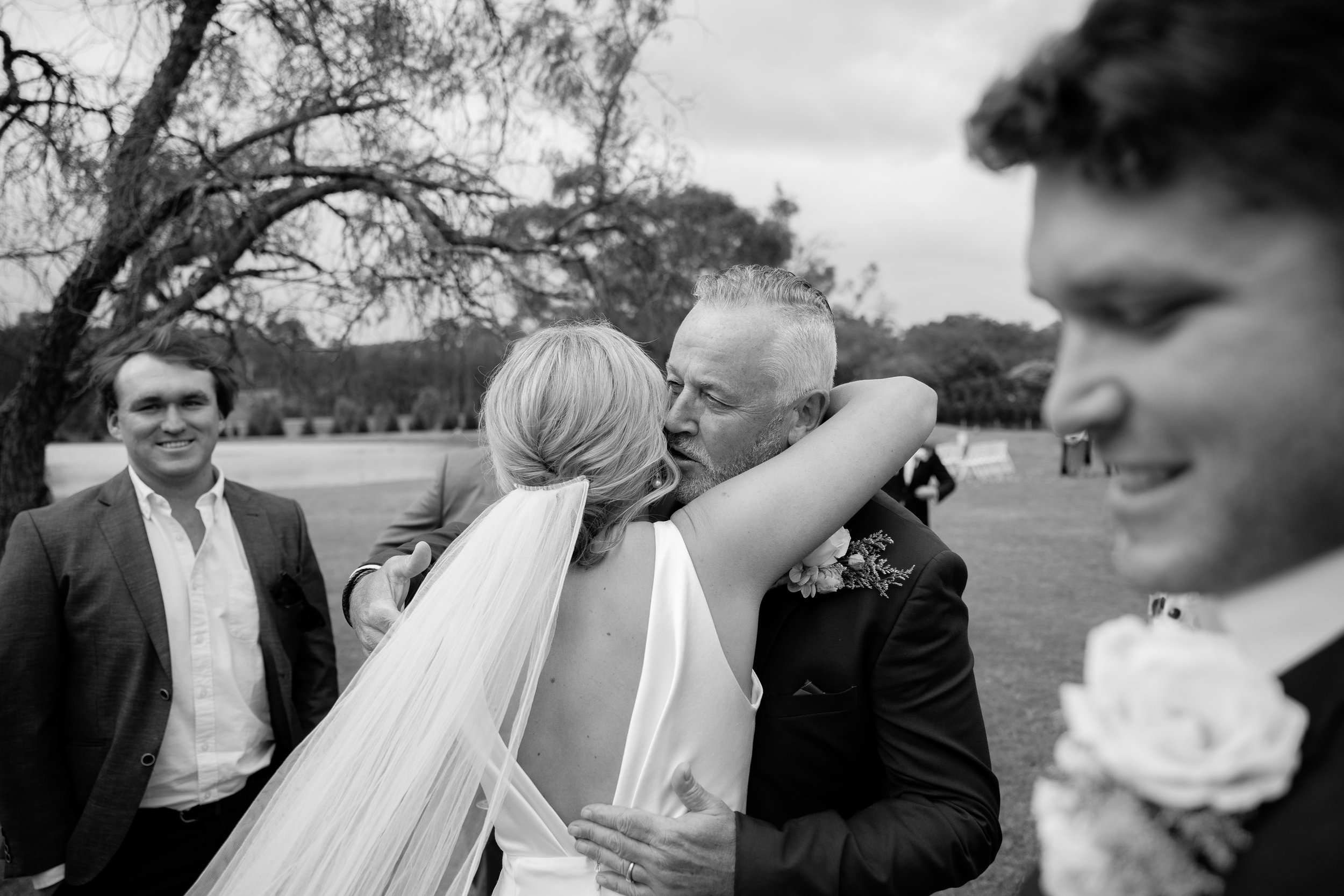 A black and white photograph of a wedding scene outdoors, featuring a bride and groom embracing and kissing, surrounded by a few guests including a man in a suit smiling, with trees and a park in the background.