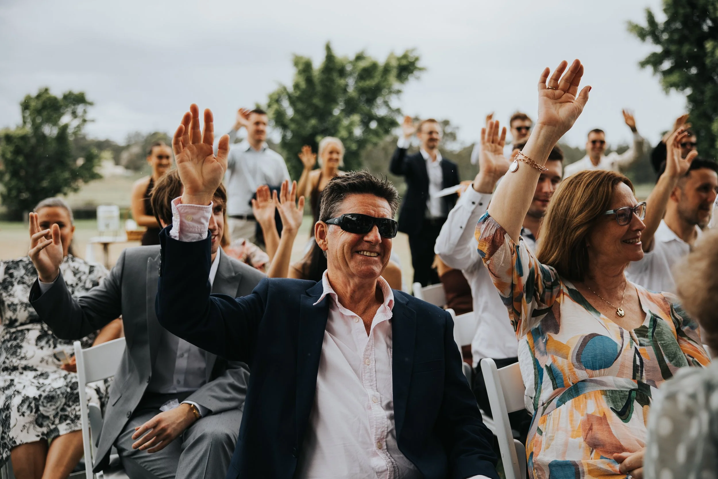 A group of people at an outdoor event raising their hands, smiling, and engaging with the speaker or activity. The scene is set in a park with trees and cloudy sky in the background.