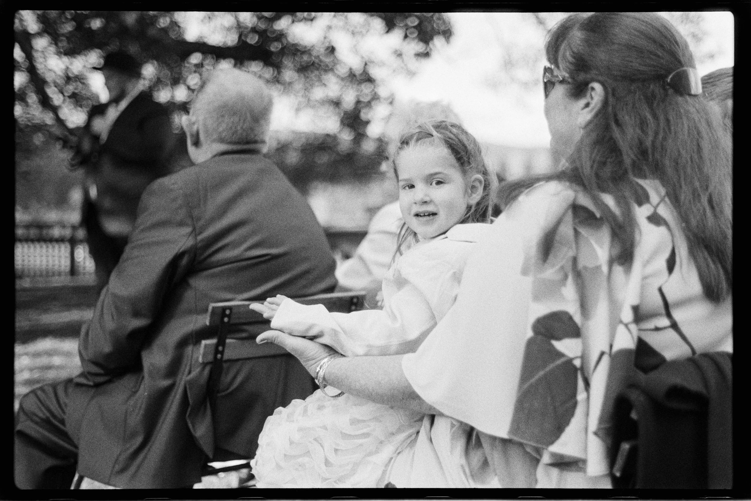 A group of children and adults sitting outdoors, engaging in conversation. One girl is smiling and looking at the camera, wearing a light-colored dress. The scene is in black and white, with blurred trees and bokeh background.