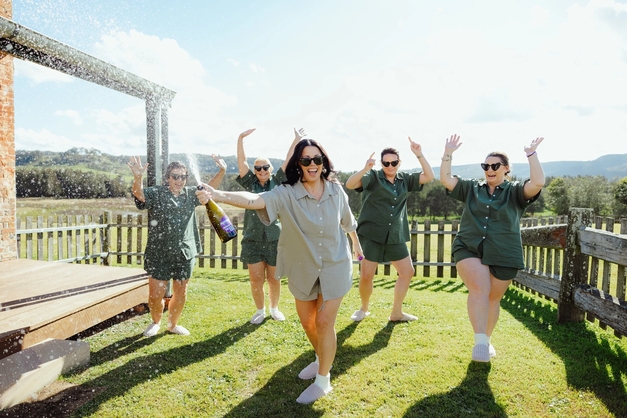 Group of women celebrating outdoors with champagne, smiling and raising their hands in the sunlight.