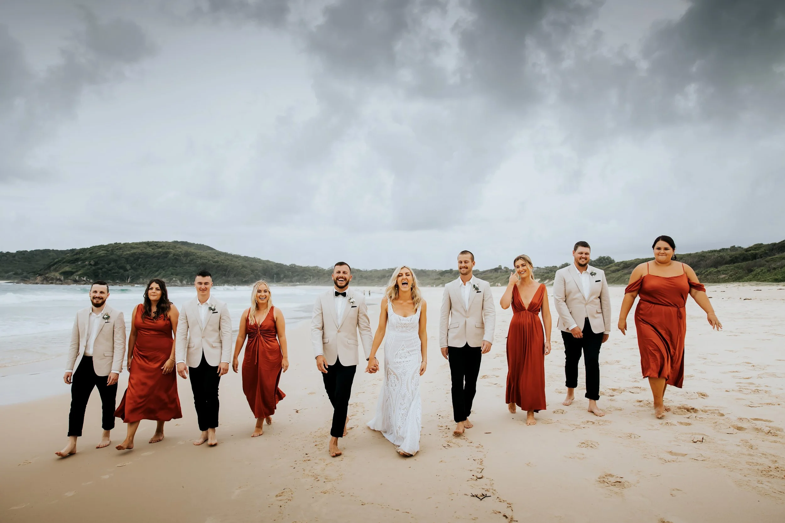 A wedding party walking along the beach, dressed in formal attire, with a cloudy sky overhead.