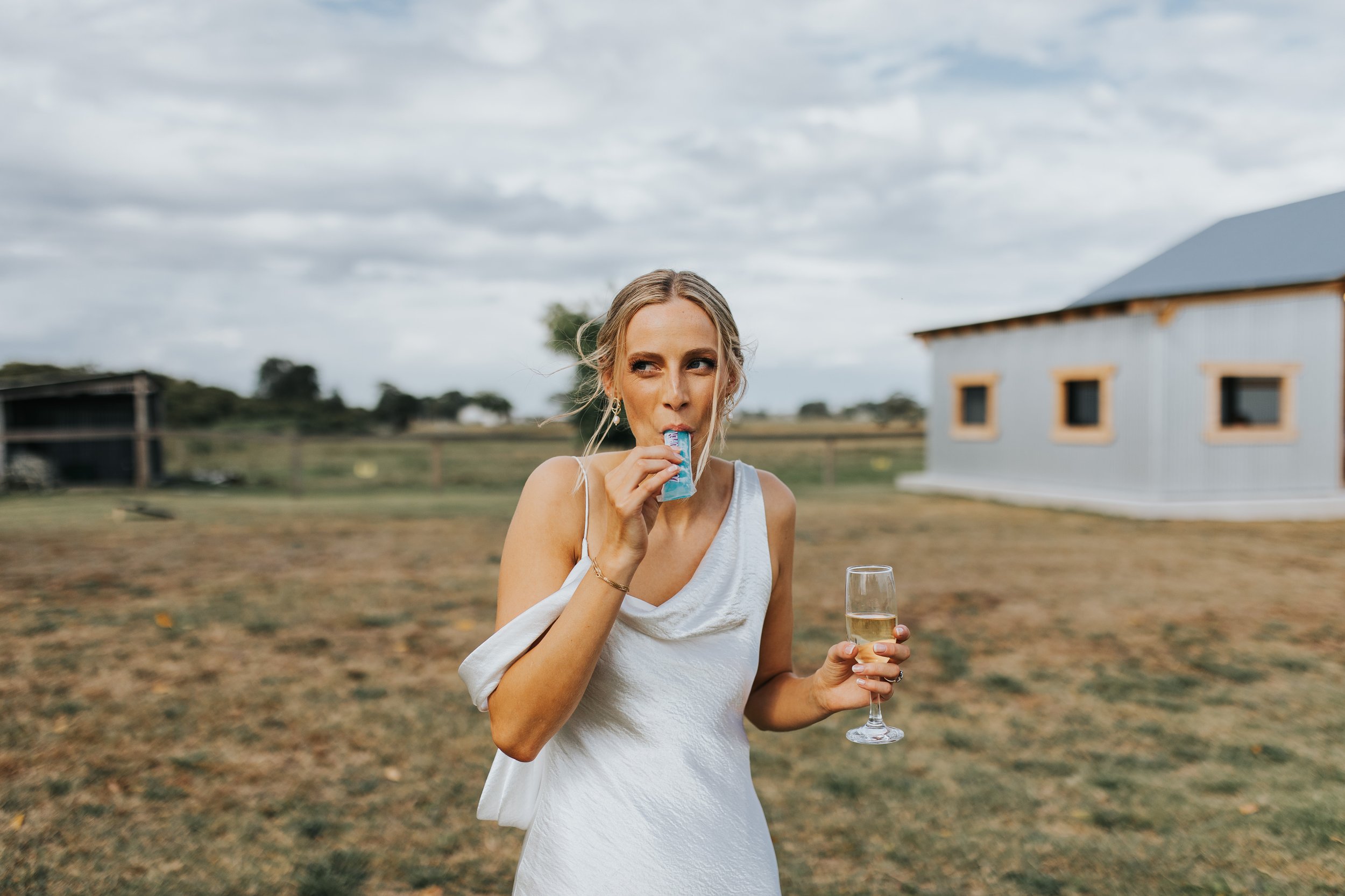 A woman in a white dress holding a glass of champagne and biting into a blue candy bar outdoors during daytime with cloudy skies.