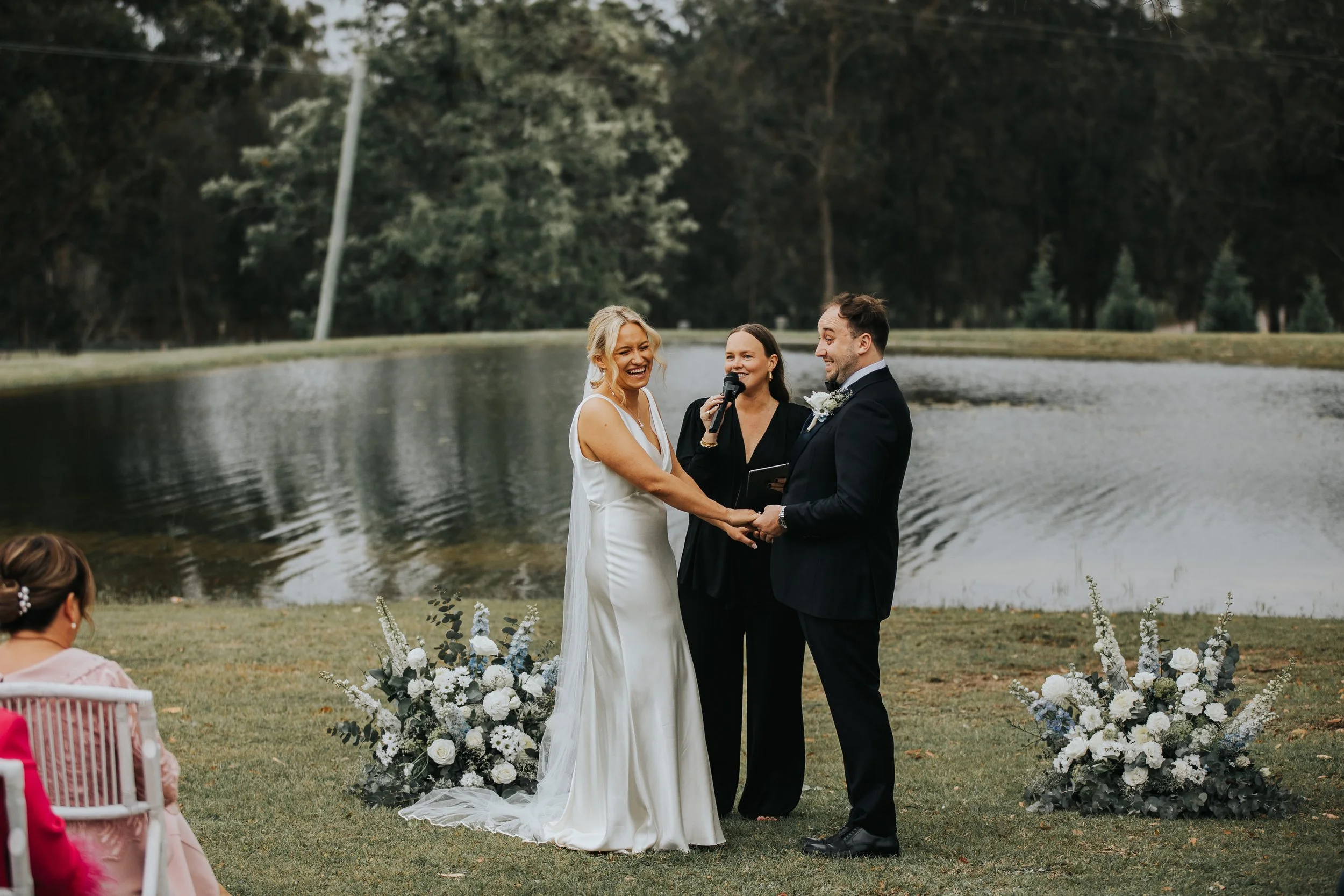 A bride and groom holding hands and smiling during a wedding ceremony outdoors by a lake, with a officiant standing between them, in front of wedding floral arrangements.