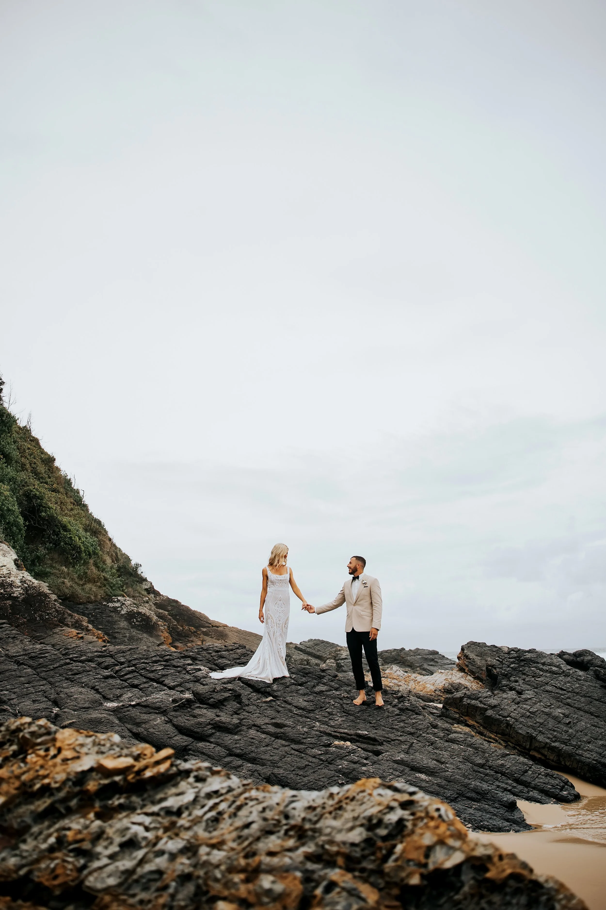 A bride and groom holding hands on a rocky beach, with the bride in a white lace wedding gown and the groom in a light-colored suit jacket and black pants, amid a cloudy sky.