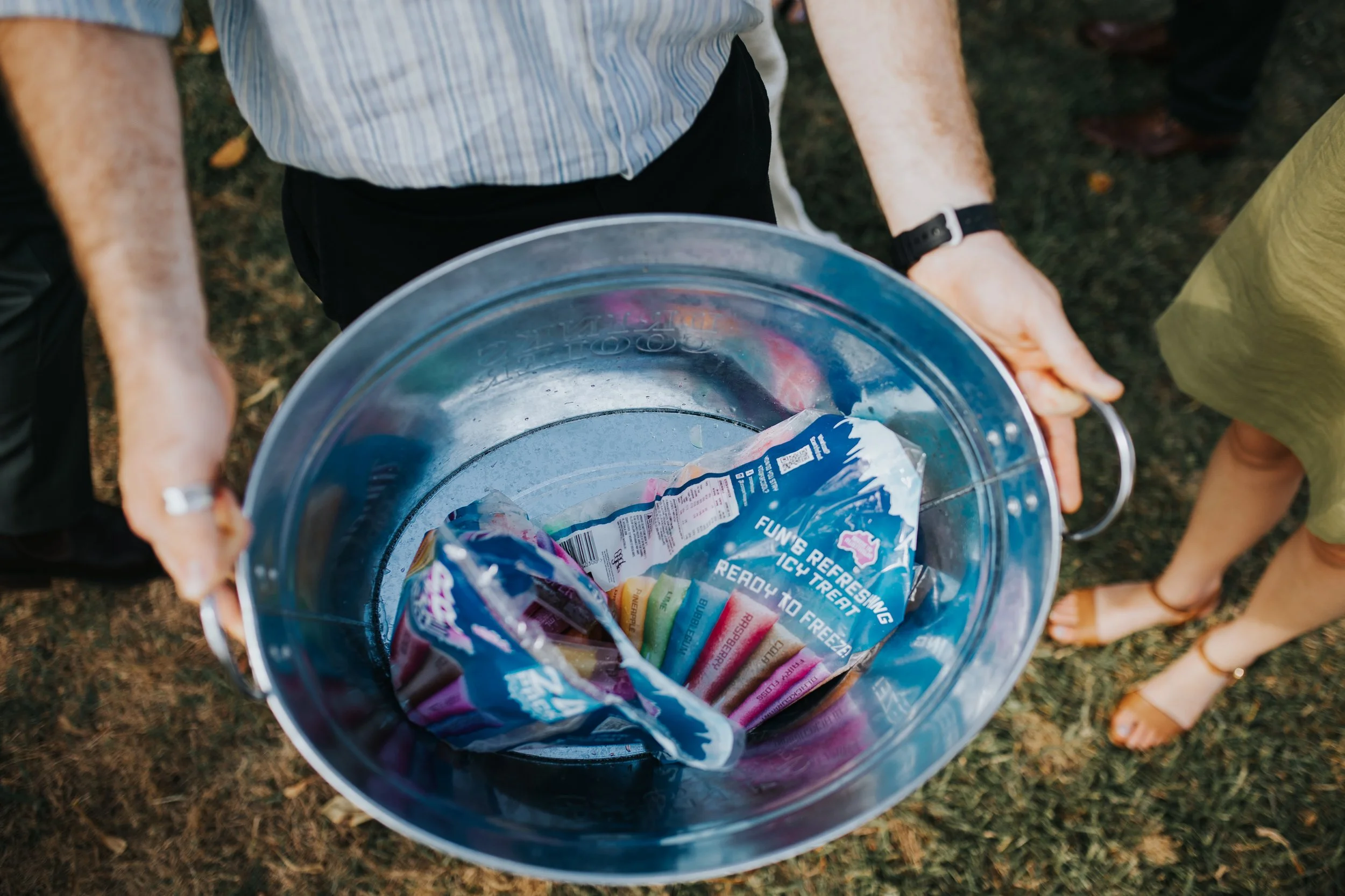 People holding a metal bucket filled with ice and assorted colorful frozen popsicles outdoors.