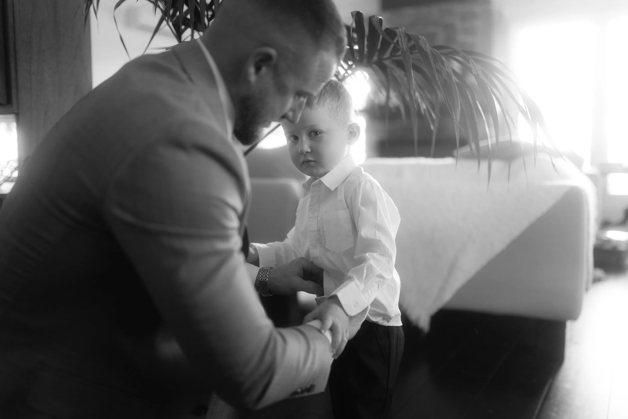 A man and a young boy are sitting at a table, engaged in a close conversation. The boy is looking directly at the camera with a serious expression, while the man appears to be focused on the boy. The setting has a warm backlight coming from the right