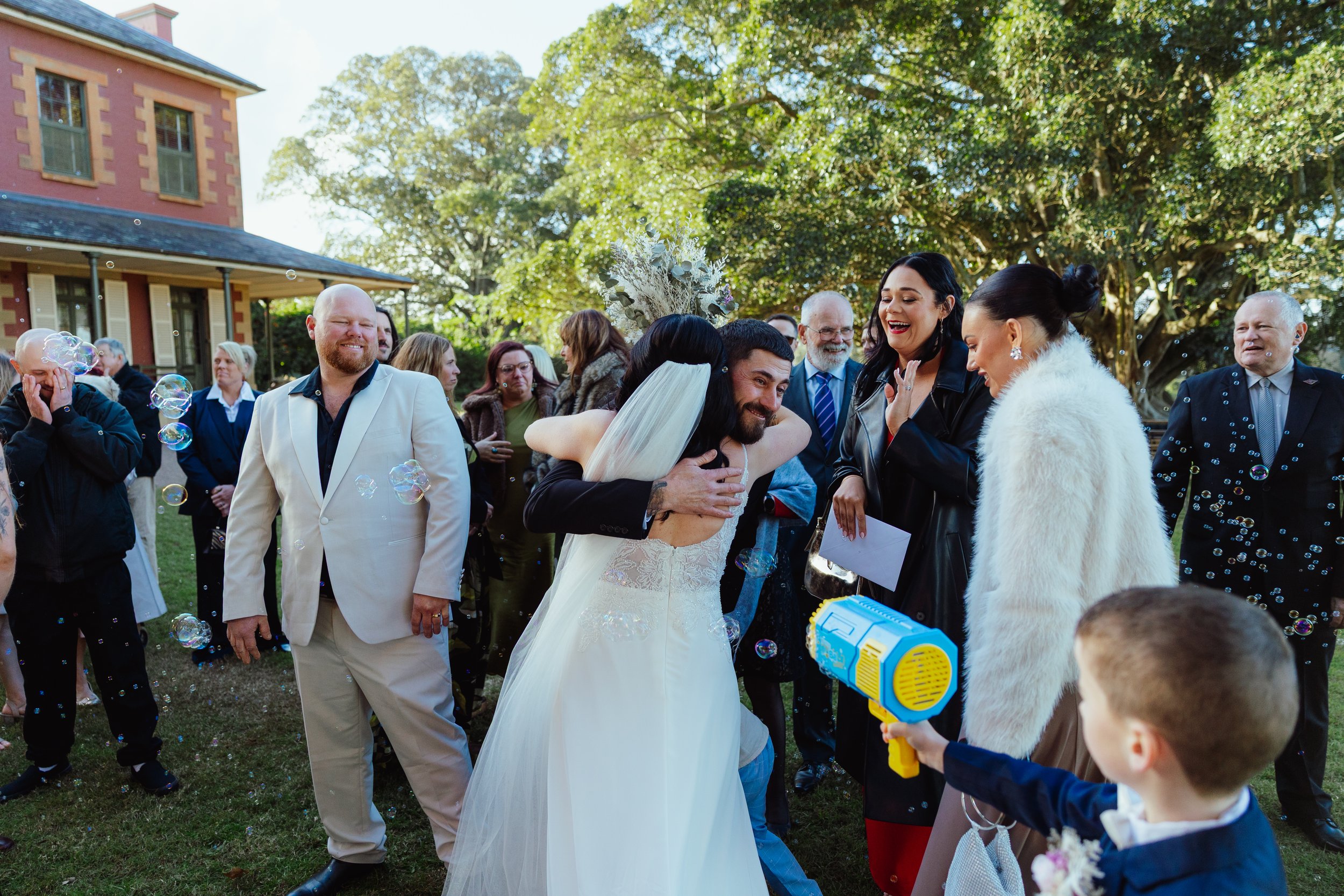 A bride and groom smiling and hugging at an outdoor wedding reception surrounded by guests, some holding bubbles and a child holding a water gun, on a green lawn with a large tree and a house in the background.