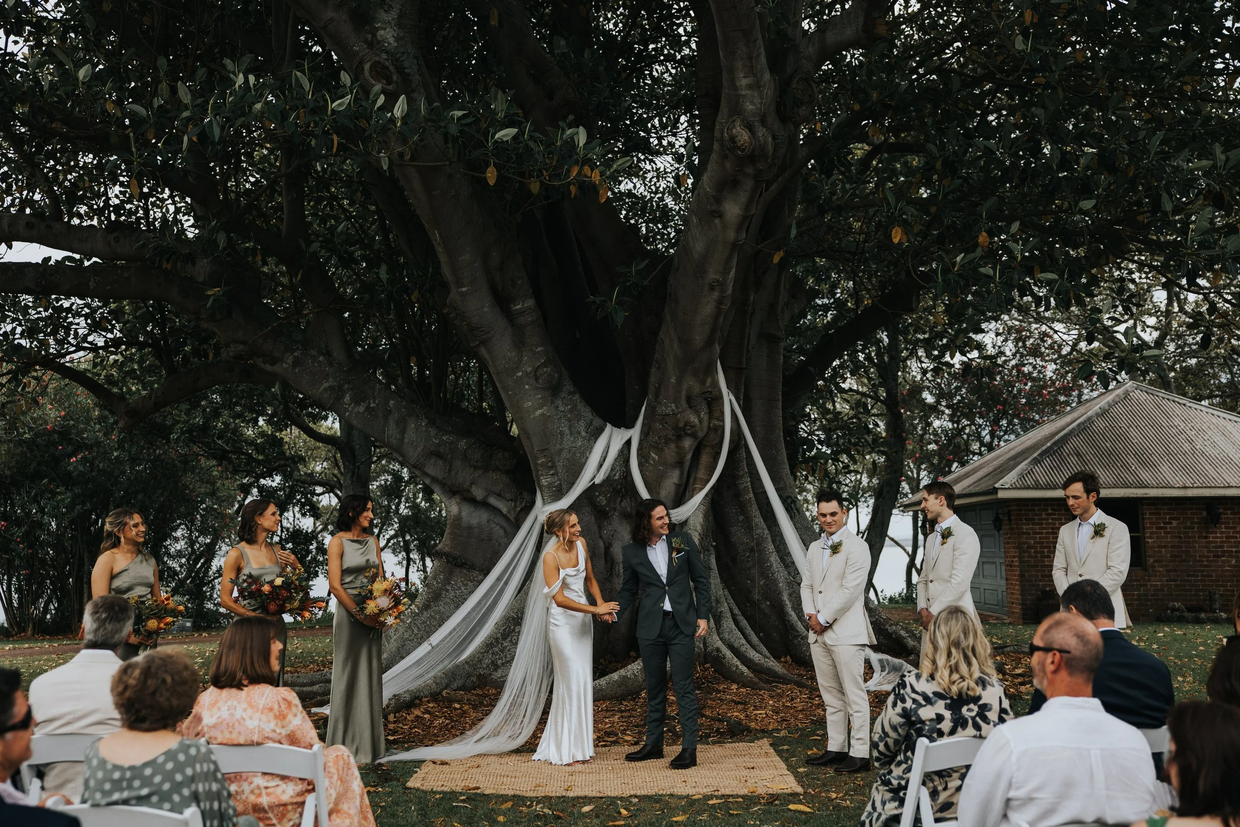 A wedding ceremony outdoors under a large tree, with the bride and groom exchanging vows, surrounded by bridesmaids, groomsmen, and seated guests.