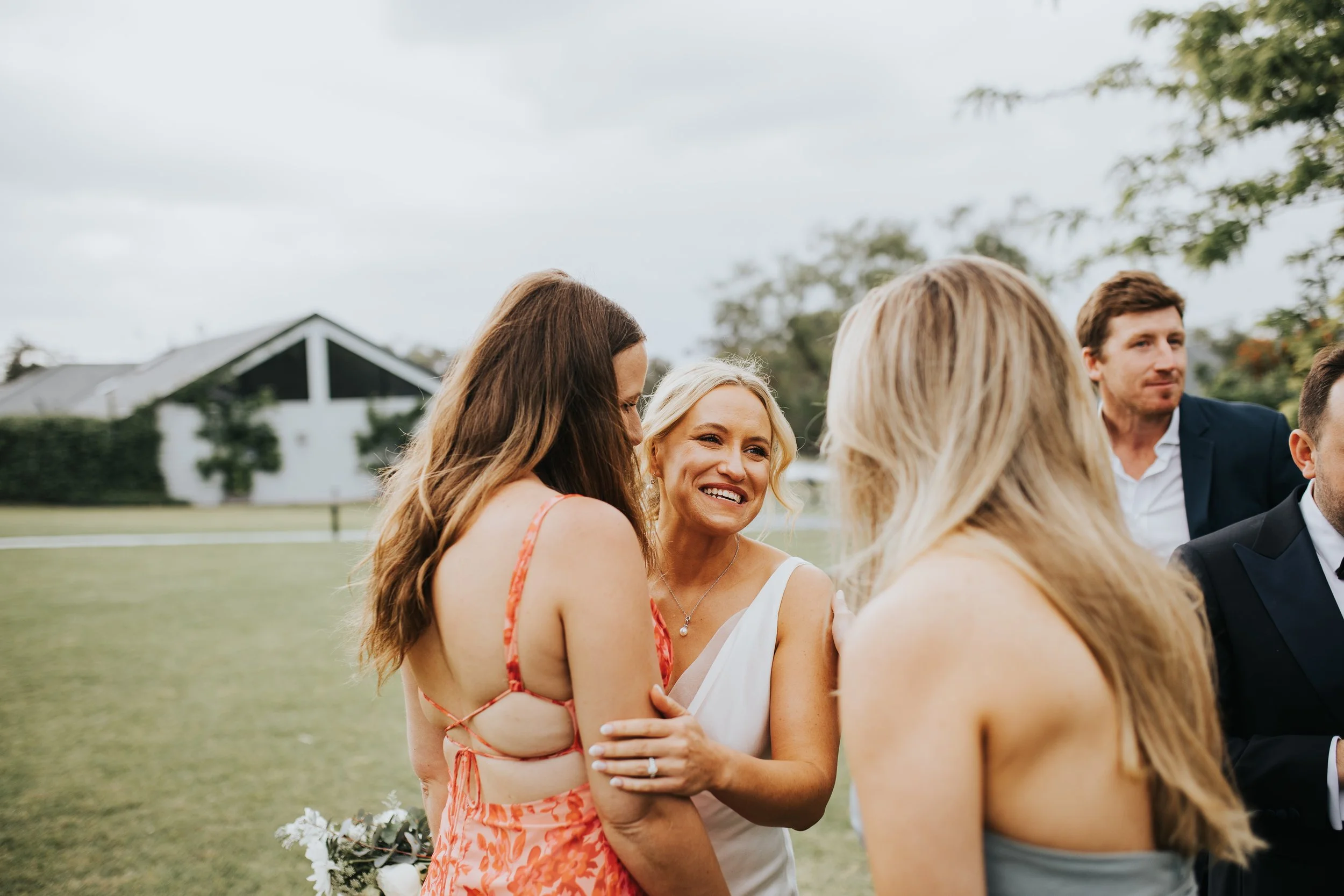 A group of people gathered outdoors on a cloudy day, with a house and trees in the background, engaged in conversation and smiling.