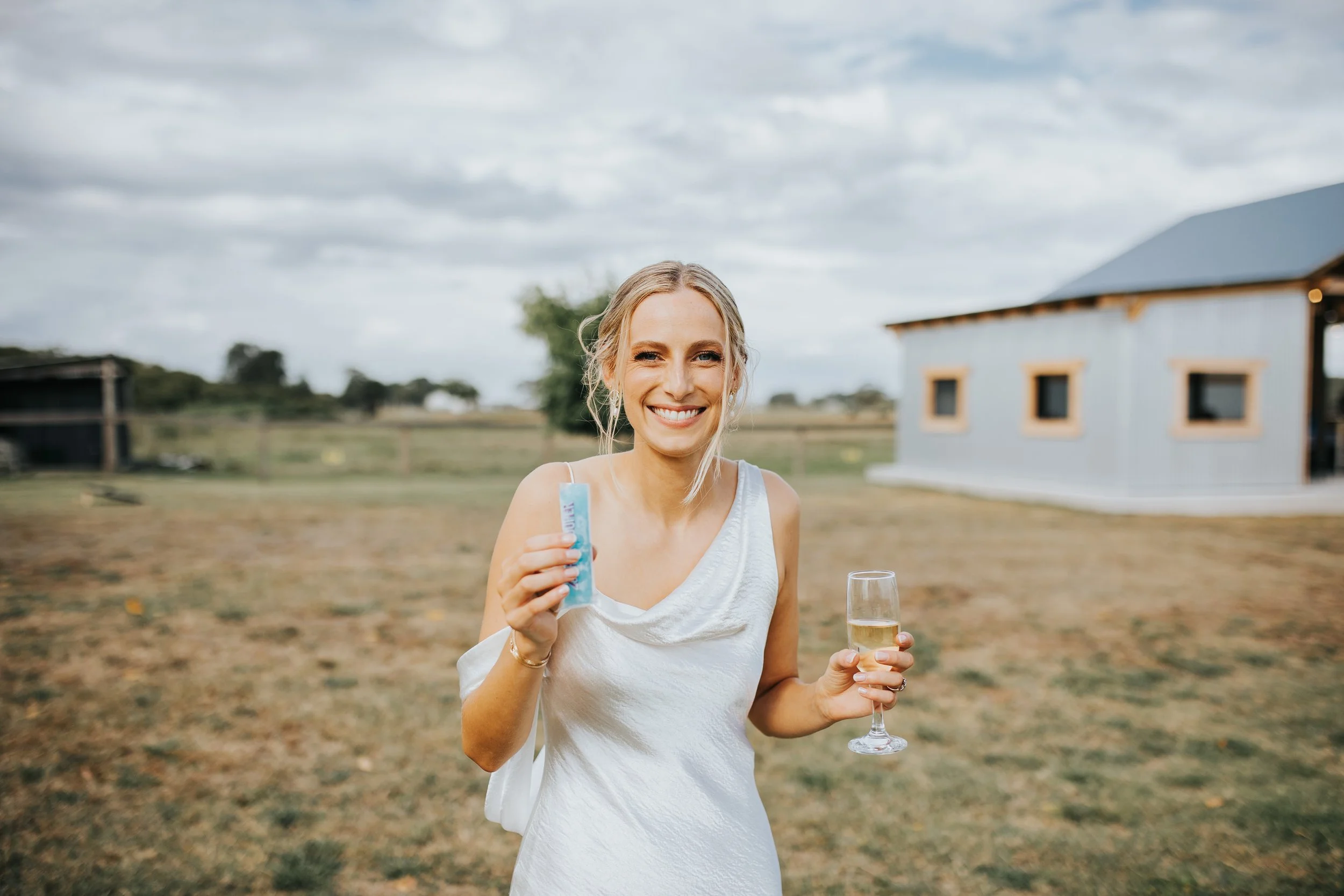 A woman smiling outdoors, holding a tube of sunscreen in one hand and a glass of champagne in the other, with a house and cloudy sky in the background.