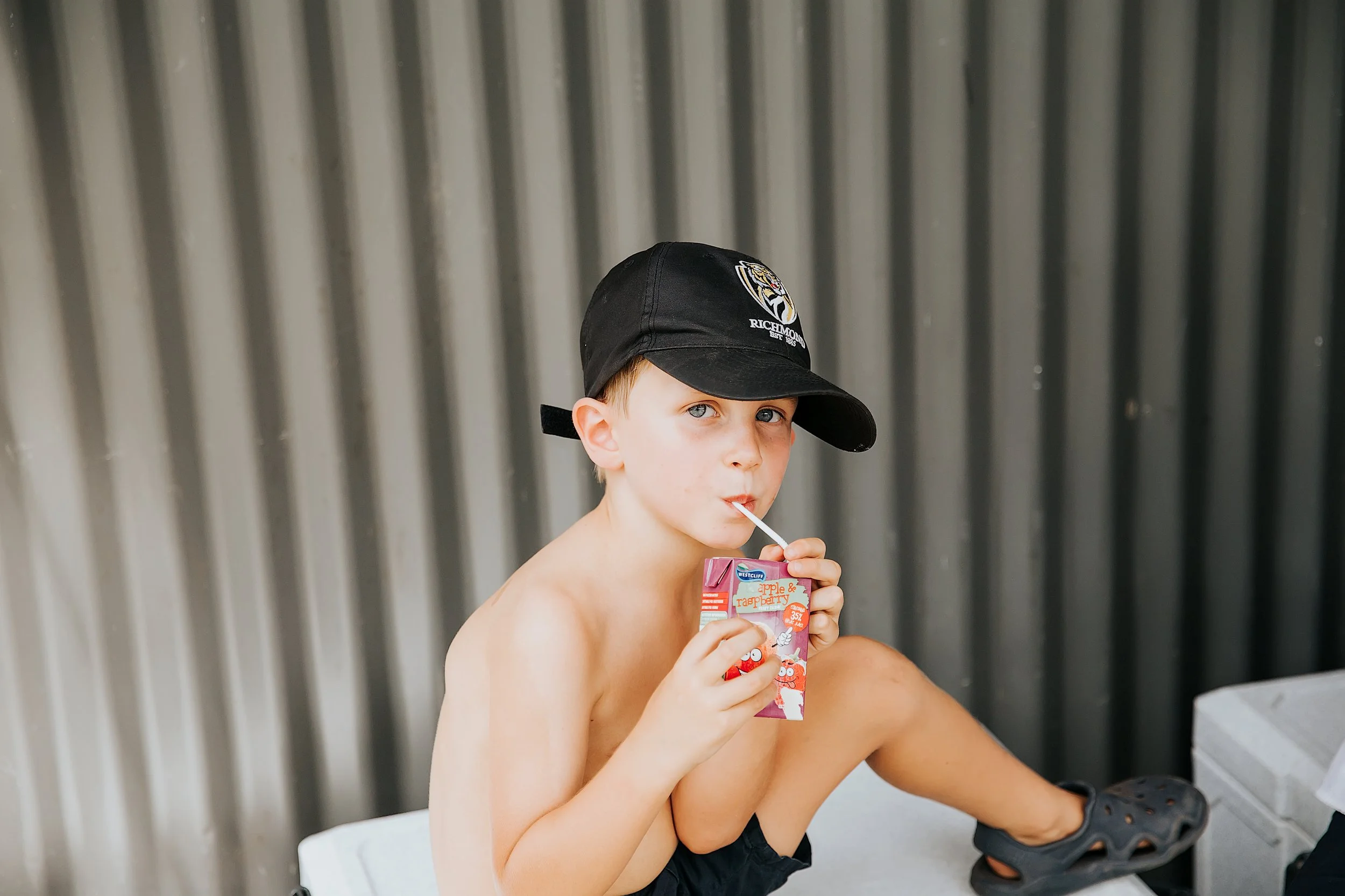 A young shirtless boy with a black cap drinking from a juice box with a straw, sitting against a corrugated metal wall.