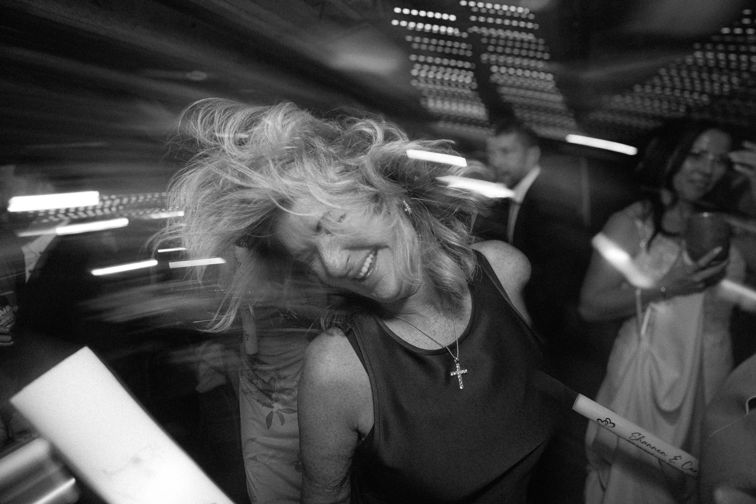 A woman with blonde curly hair and a cross necklace smiling, with two men in the background, in a dimly lit indoor setting.