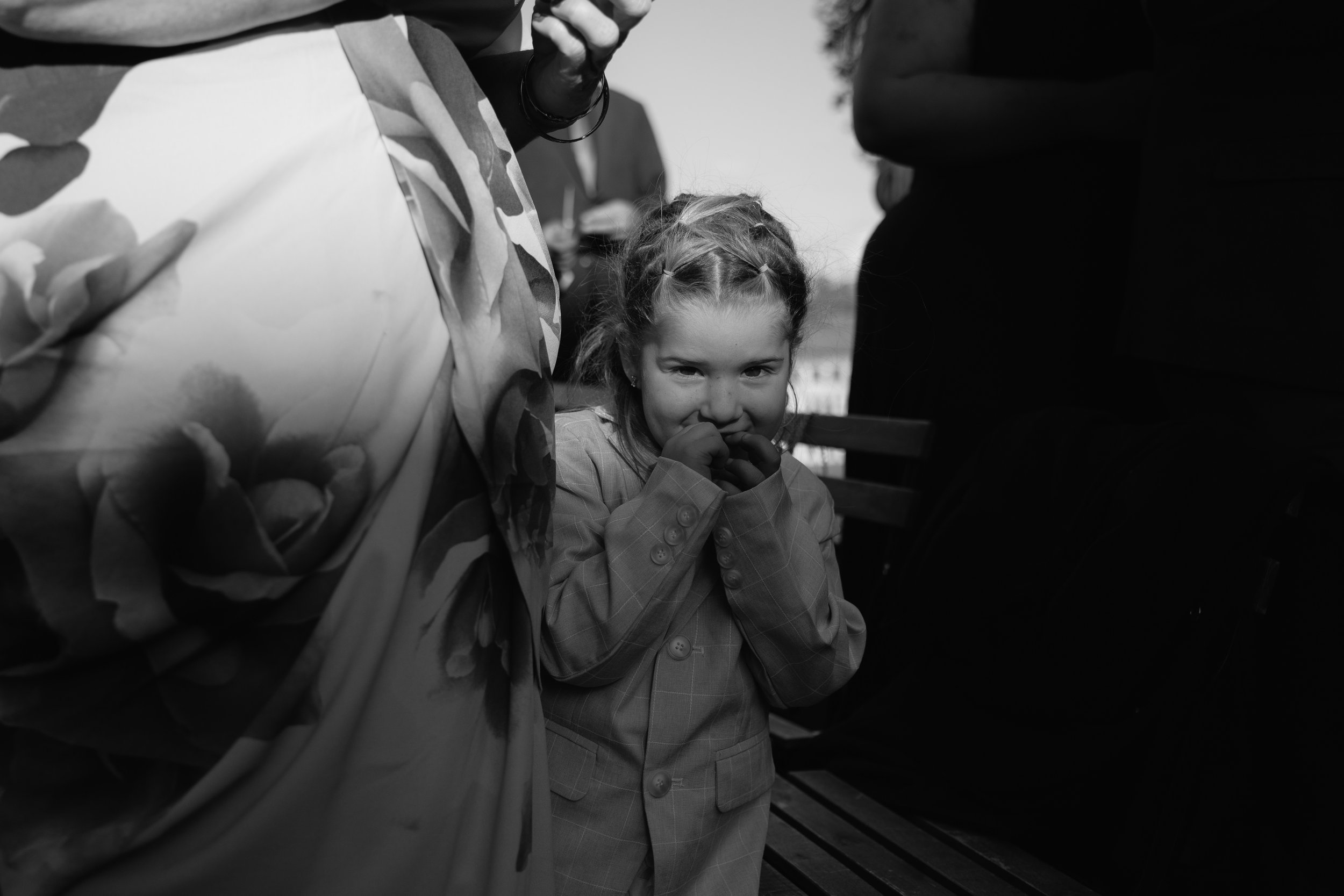 A young girl with braided hair wearing a checked blazer is covering her mouth with her hands while looking towards the camera. Two adults are partially visible next to her, one in a patterned jacket and the other in dark clothing, in an outdoor setti