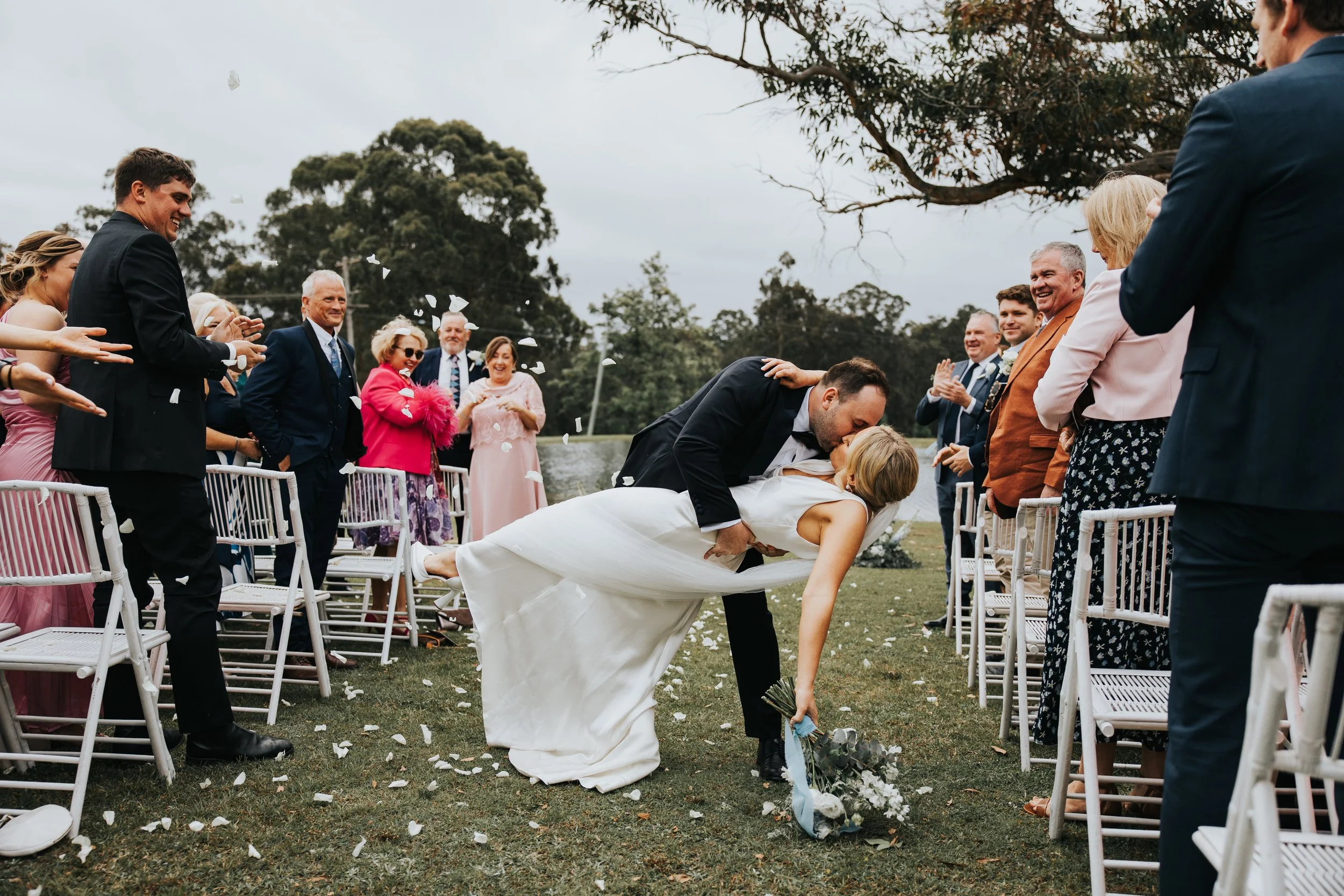 A bride and groom kissing during their outdoor wedding ceremony, with guests celebrating and throwing flower petals on a grassy area under cloudy skies.